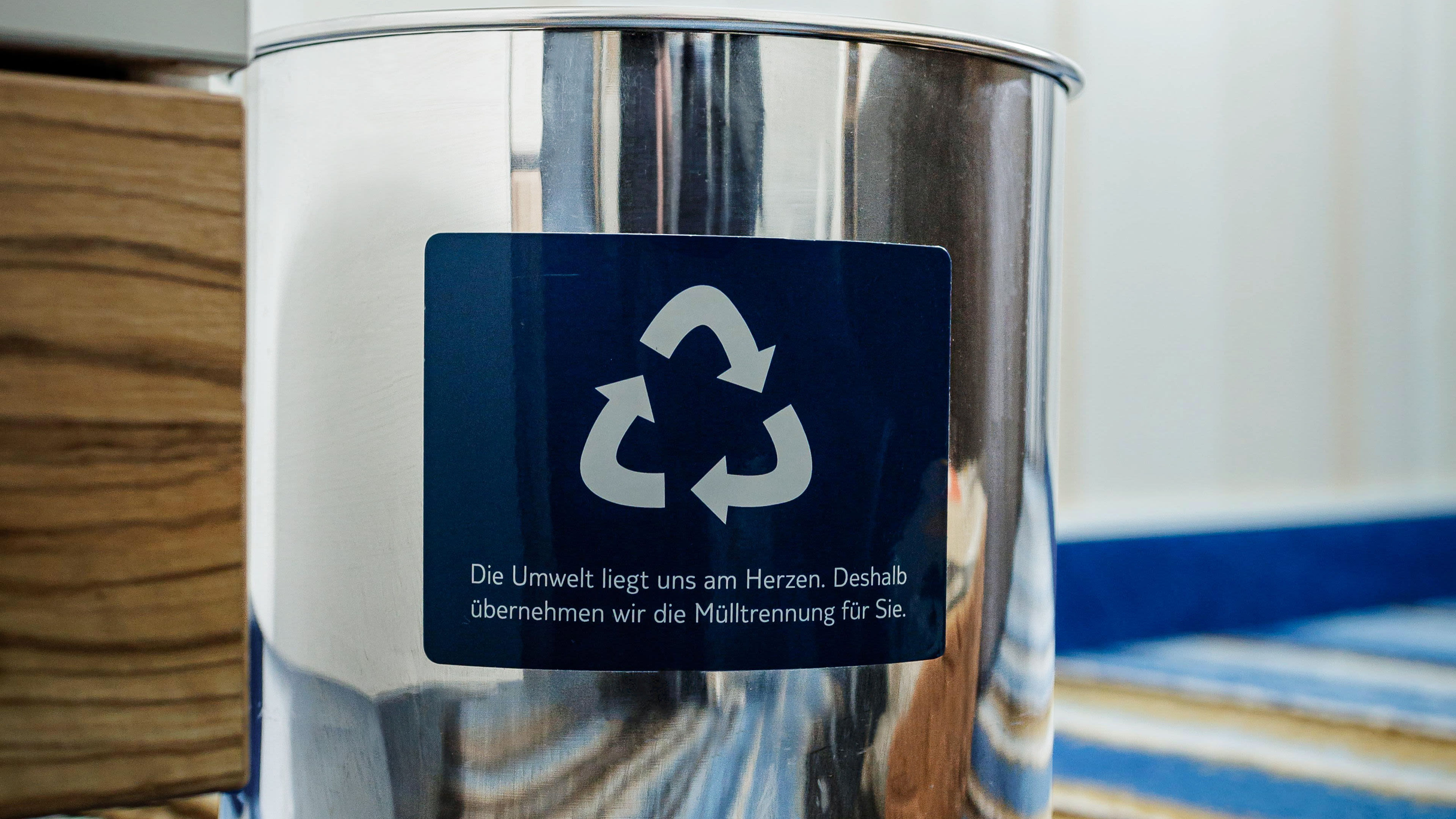 Recycling-Symbol auf silbernem Mülleimer mit Holz- und Teppichhintergrund.