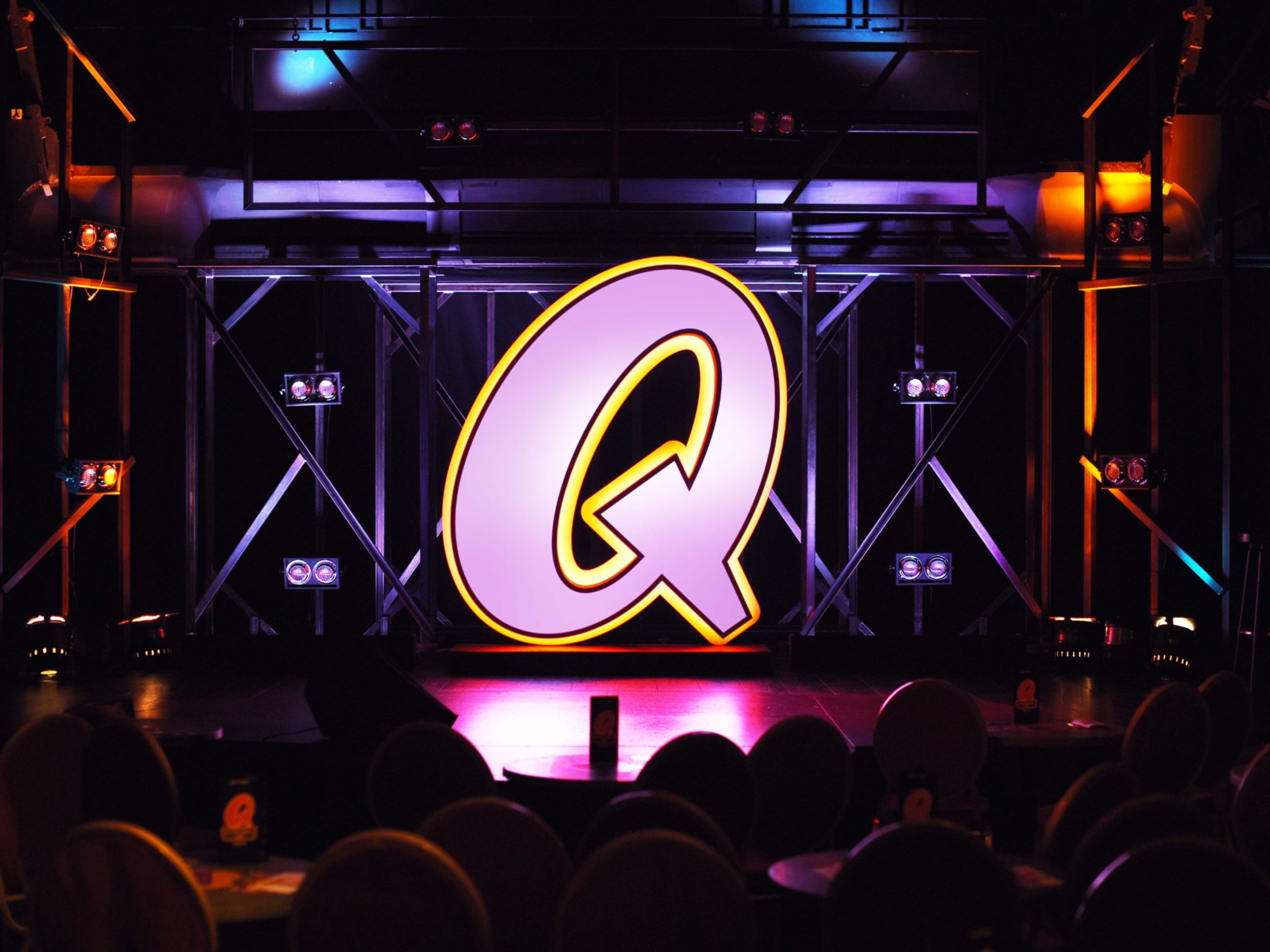 Quatsch Comedy Bühnenfoto Großes leuchtendes "Q" auf Bühne, bunte Lichter, dunkler Hintergrund.