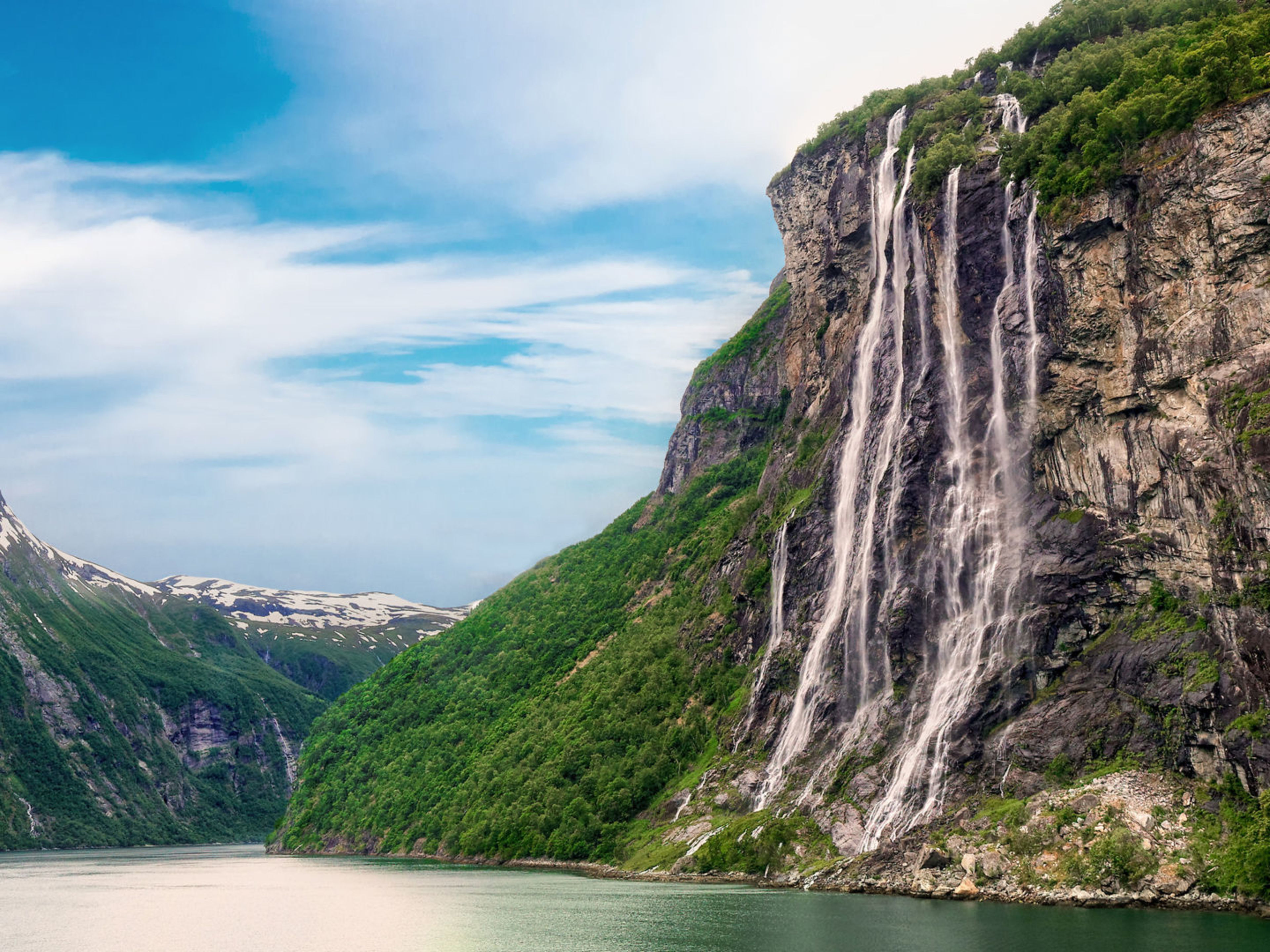 Geiranger Norwegen Wasserfall im Geirangerfjord, Norwegen, mit grünen Bergen und blauem Himmel.