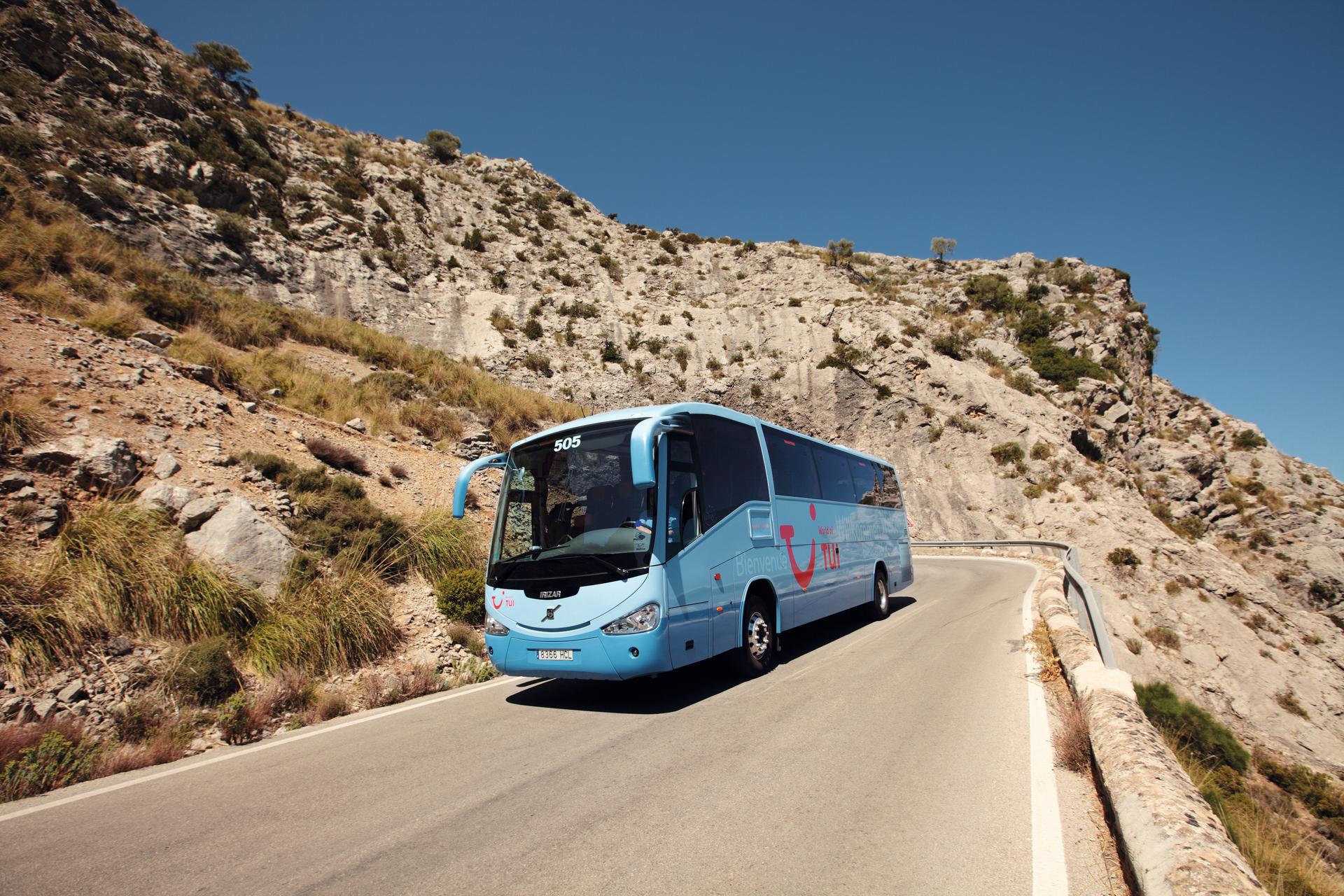 Blauer Reisebus auf kurviger Bergstraße bei Sonnenschein.