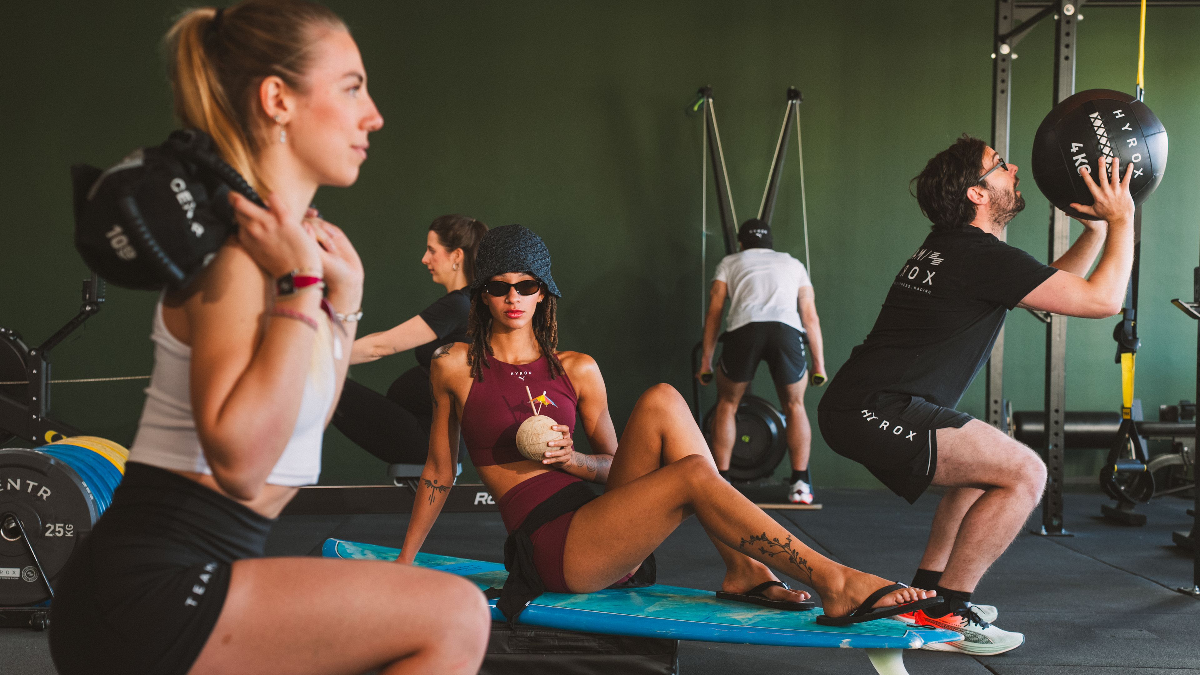 HYROX Cruise (2) Menschen trainieren im Fitnessstudio, Frau sitzt mit Kokosnuss.