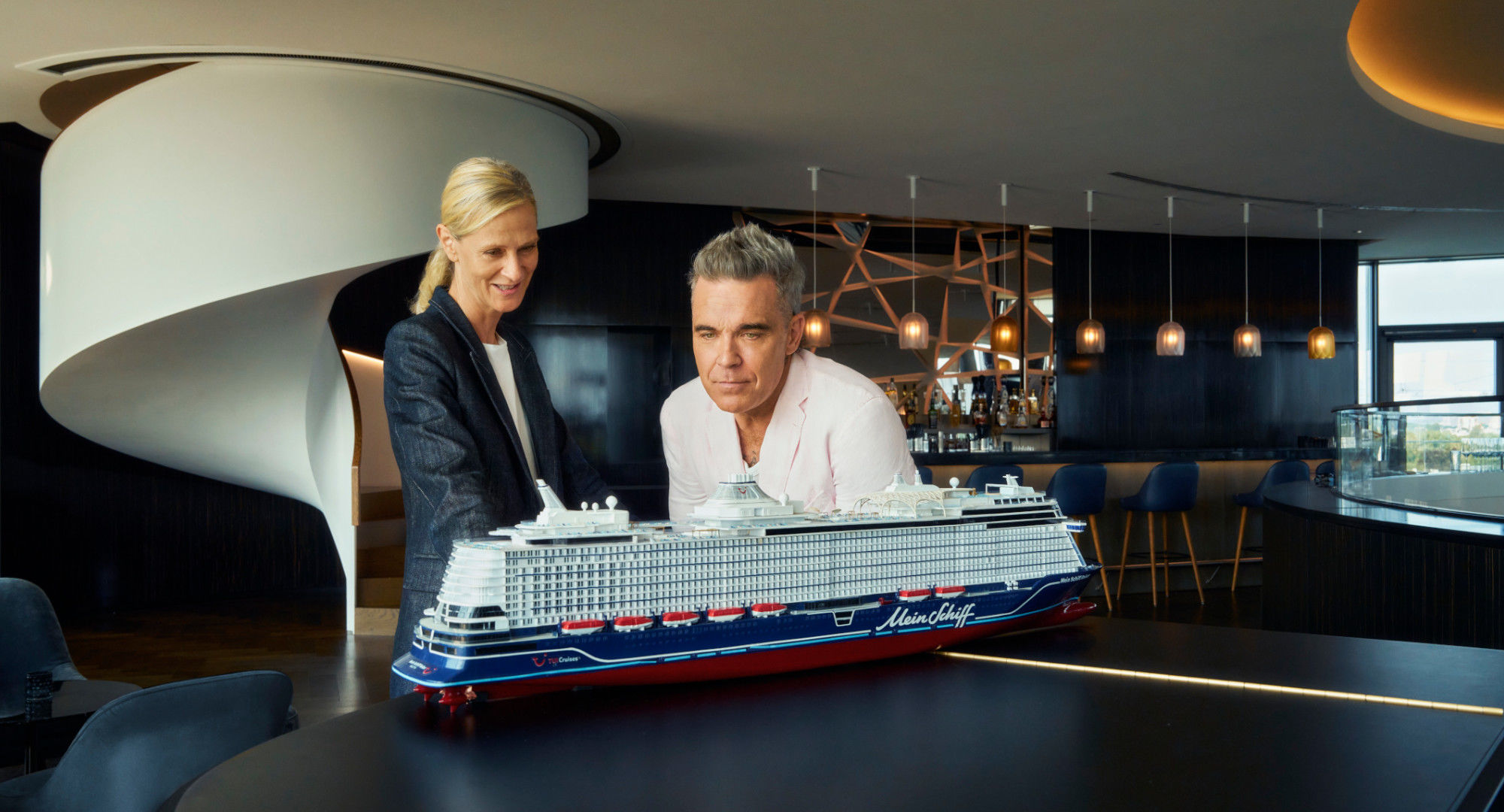 Zwei Personen betrachten Modell eines Kreuzfahrtschiffs der Mein Schiff® Flotte in modernem Raum.