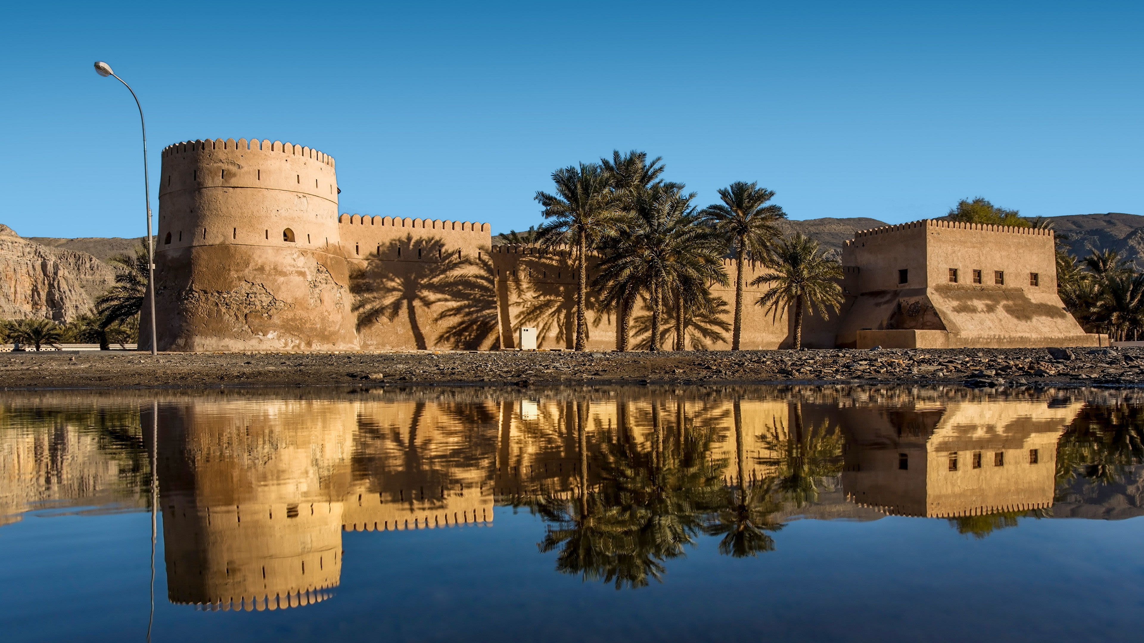 Kahsab Oman Altes Fort mit Palmen und Spiegelung im Wasser, blauer Himmel.