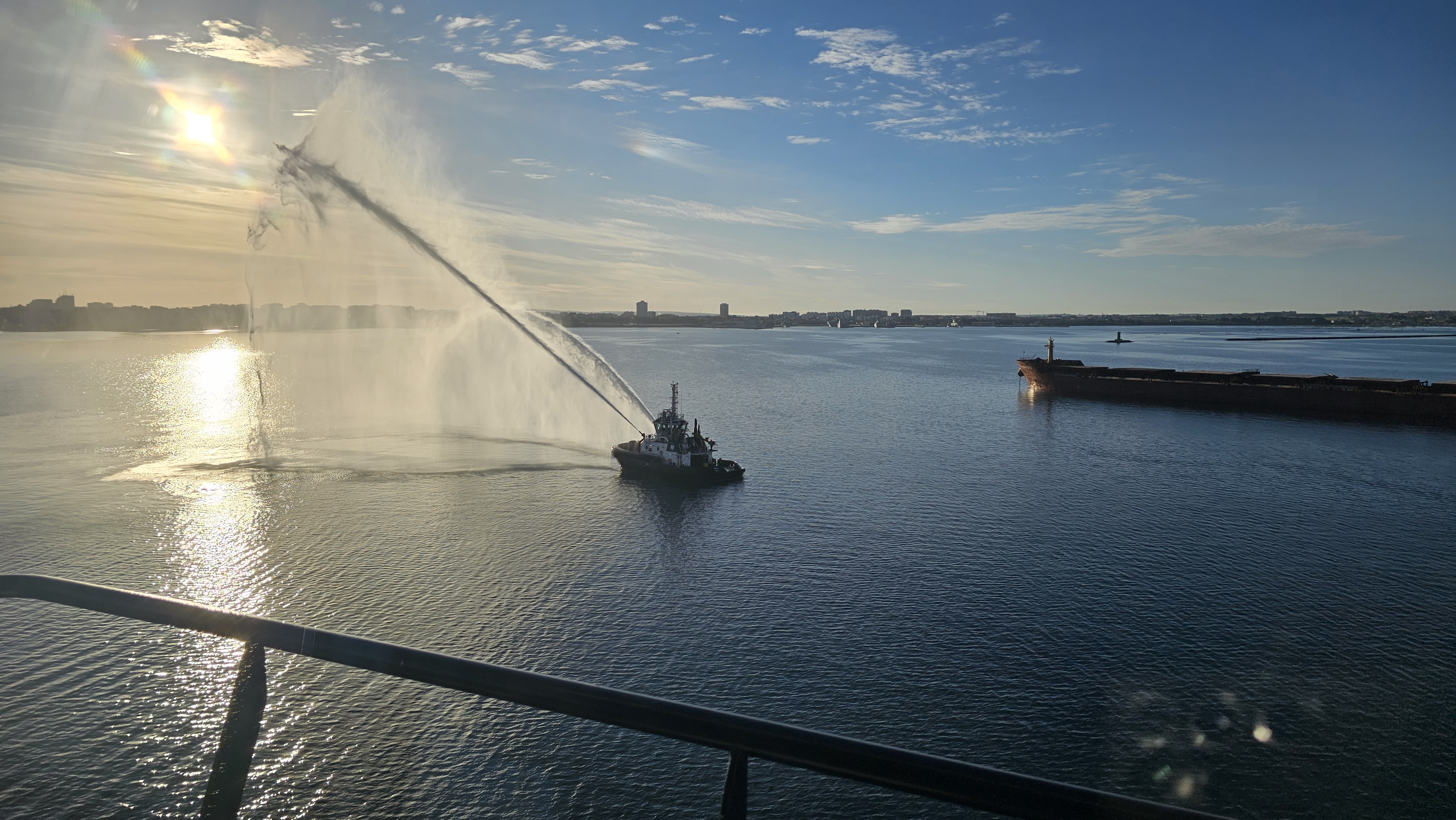 Schlepper sprüht Wasser im Hafen bei Sonnenuntergang, Frachtschiff im Hintergrund.