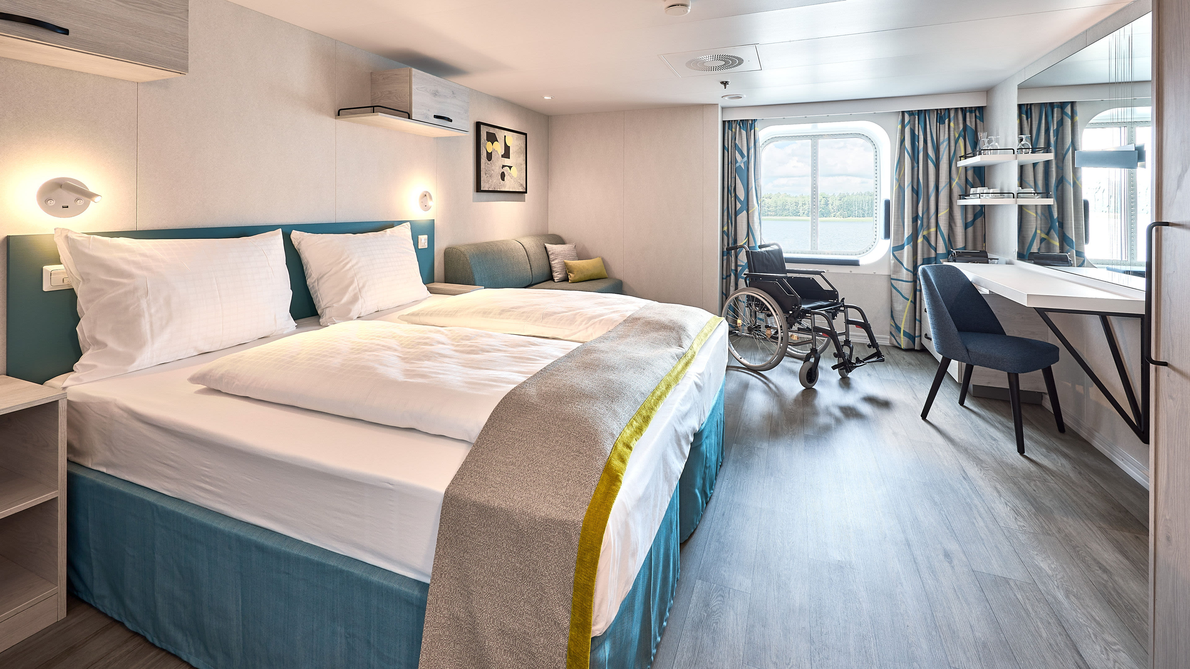 Barrierefreies Kreuzfahrtschiffzimmer mit Doppelbett, Rollstuhl, Schreibtisch und Fensterblick.