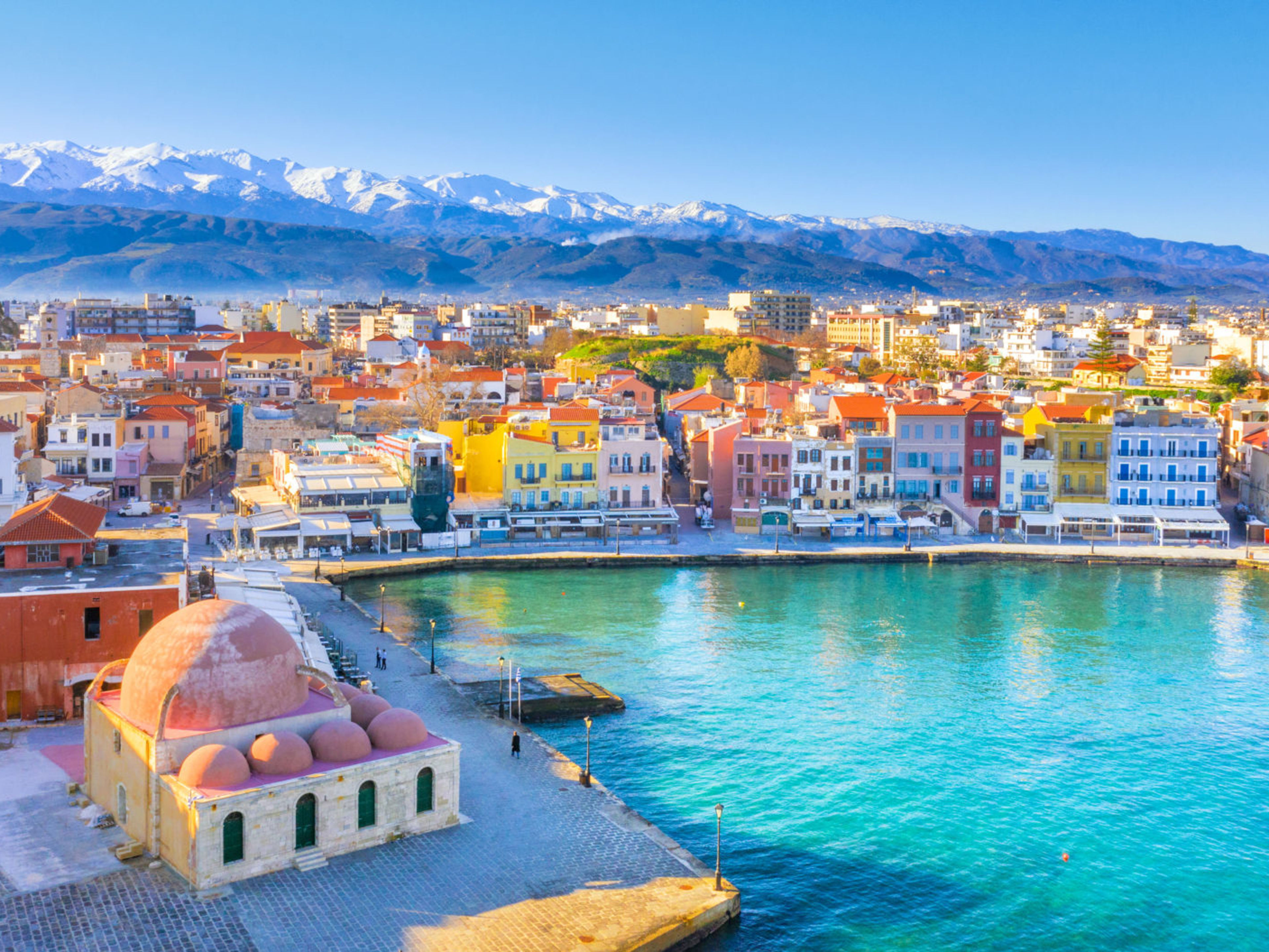 Hafenbild - Souda Chania Kreta Bunte Häuser am Hafen von Chania, Kreta, mit schneebedeckten Bergen im Hintergrund.