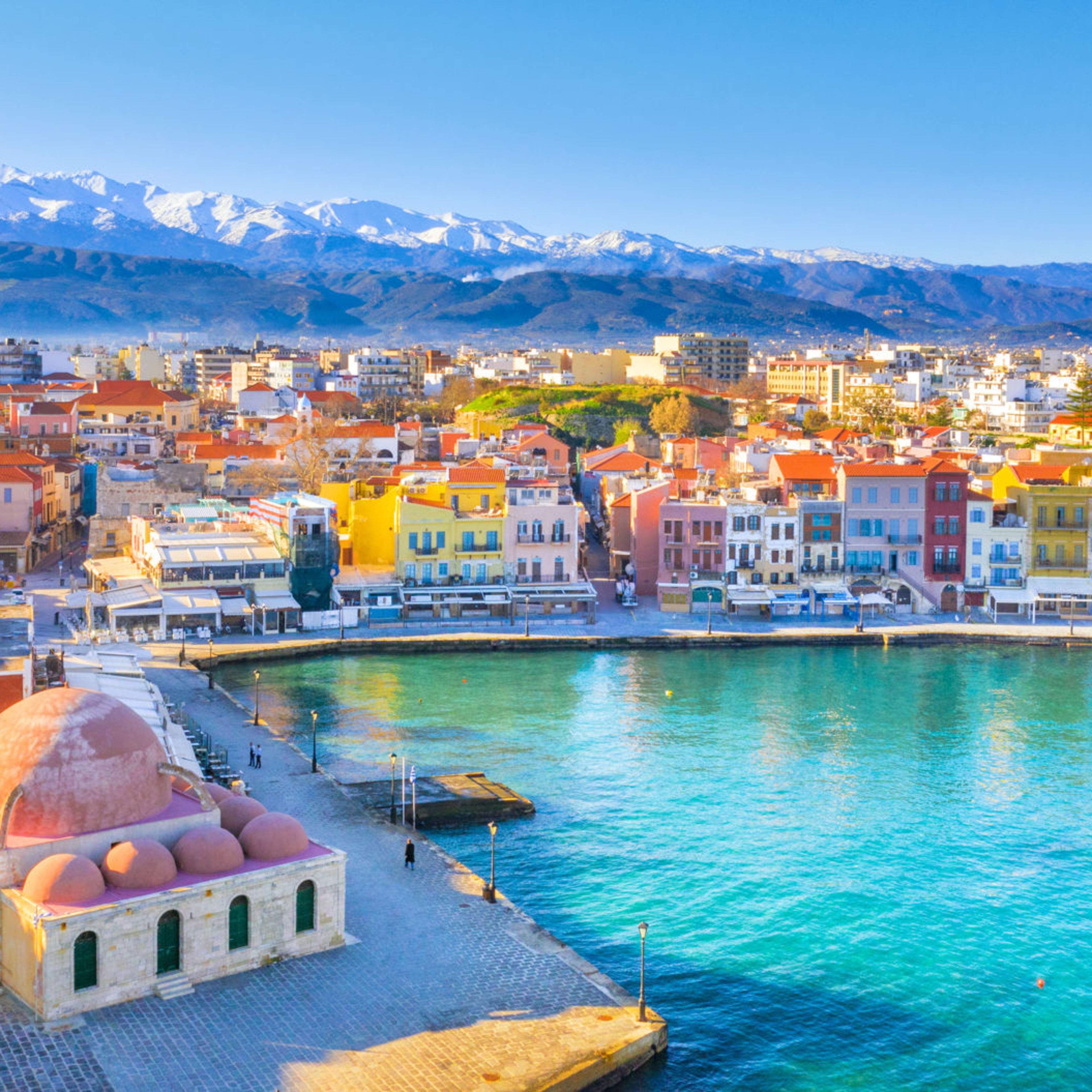 Hafenbild - Souda Chania Kreta Bunte Häuser am Hafen von Chania, Kreta, mit schneebedeckten Bergen im Hintergrund.