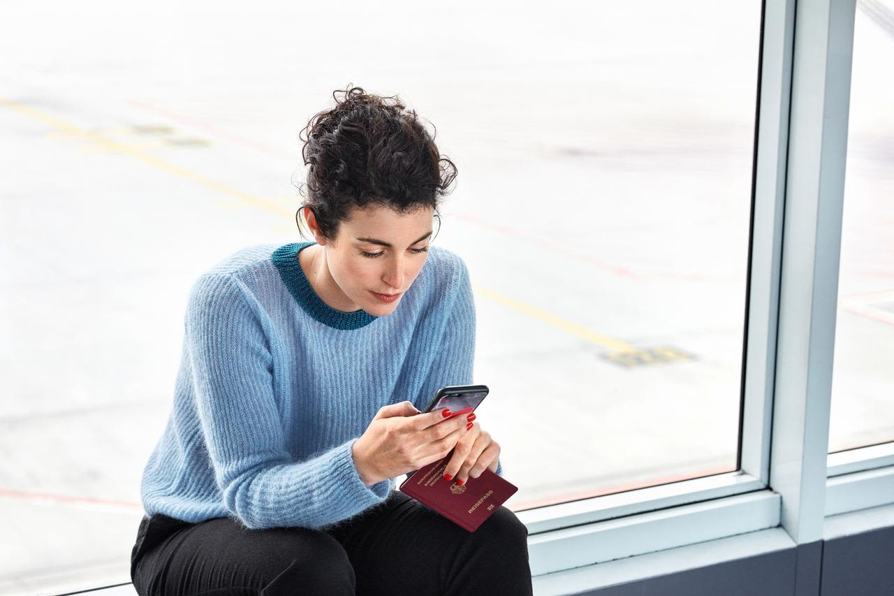 Frau im blauen Pullover am Flughafen mit Smartphone und Reisepass.