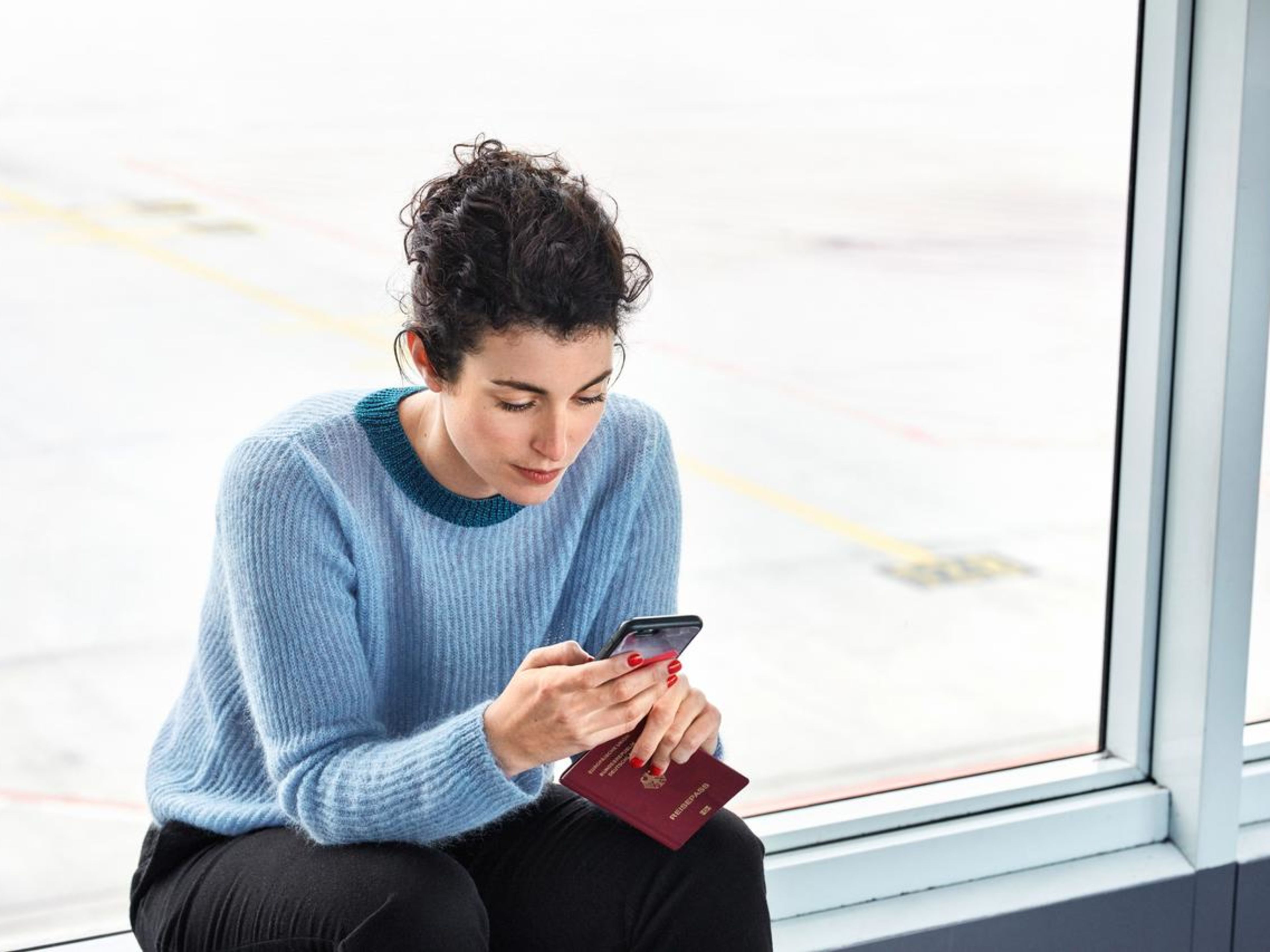 Frau im blauen Pullover am Flughafen mit Smartphone und Reisepass. Frau im blauen Pullover am Flughafen mit Smartphone und Reisepass.