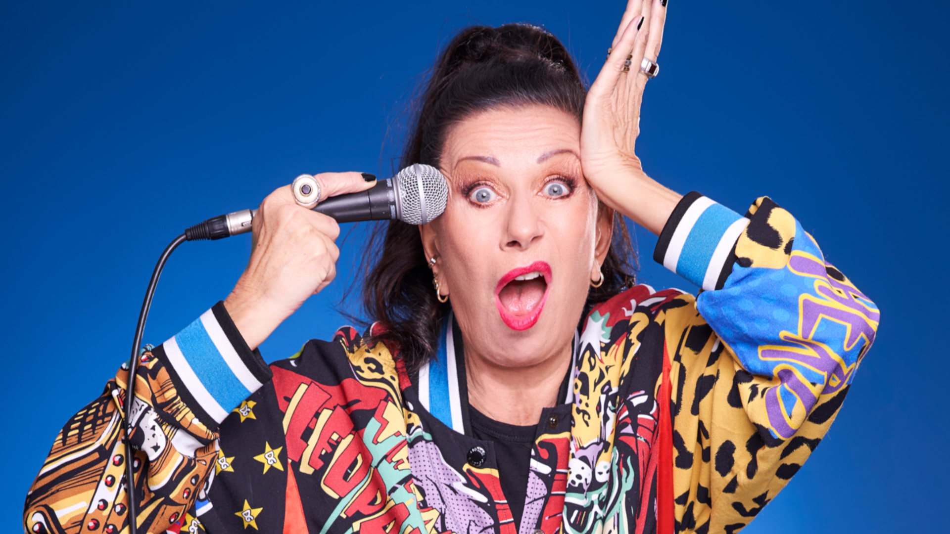 Frau mit Mikrofon, bunter Pullover, überrascht, blauer Hintergrund.