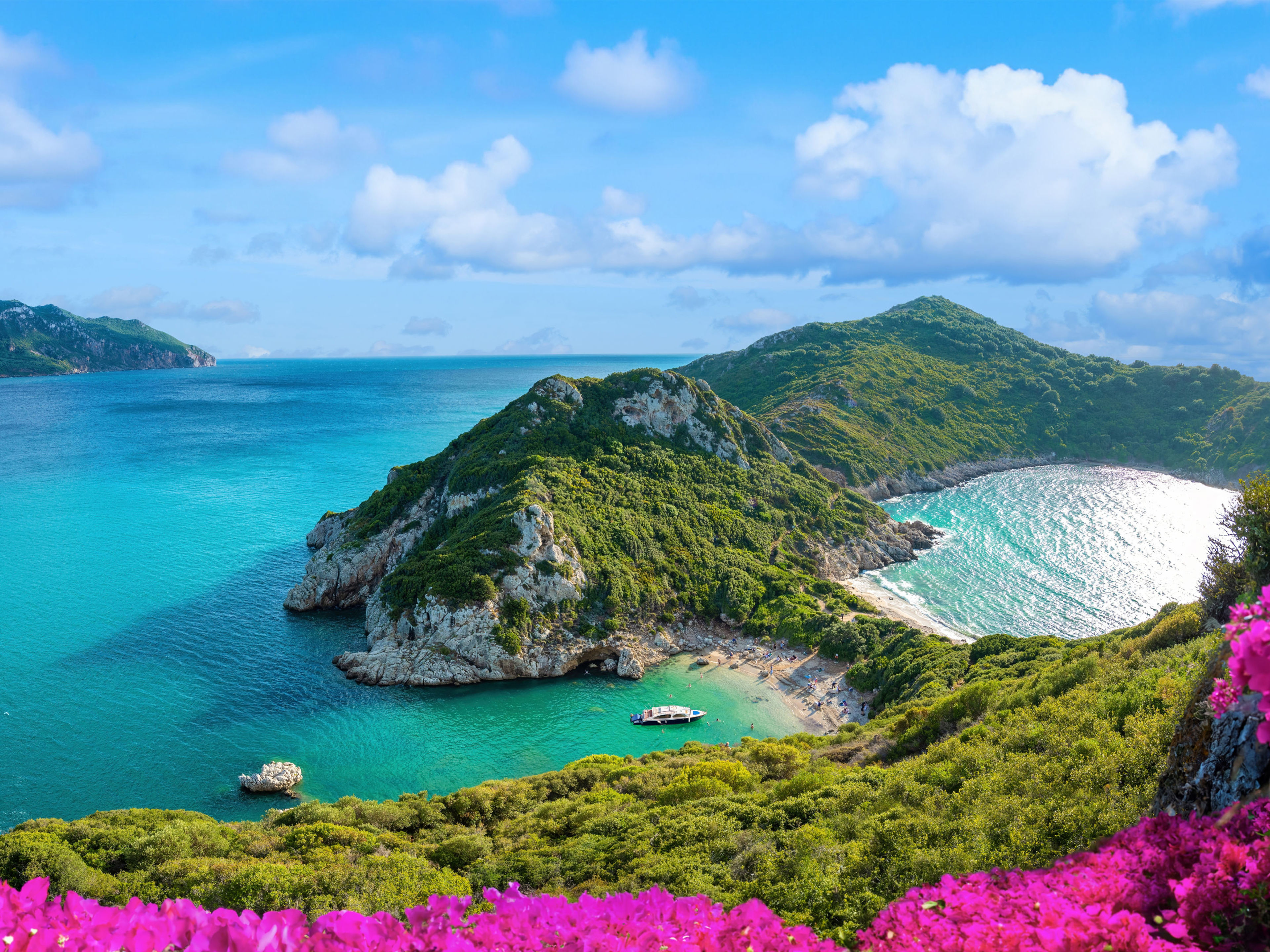 Erleben Sie Korfu bei Ihrer Mittelmeer-Kreuzfahrt Küste von Korfu mit türkisfarbenem Meer, grünen Hügeln und pinken Blumen.