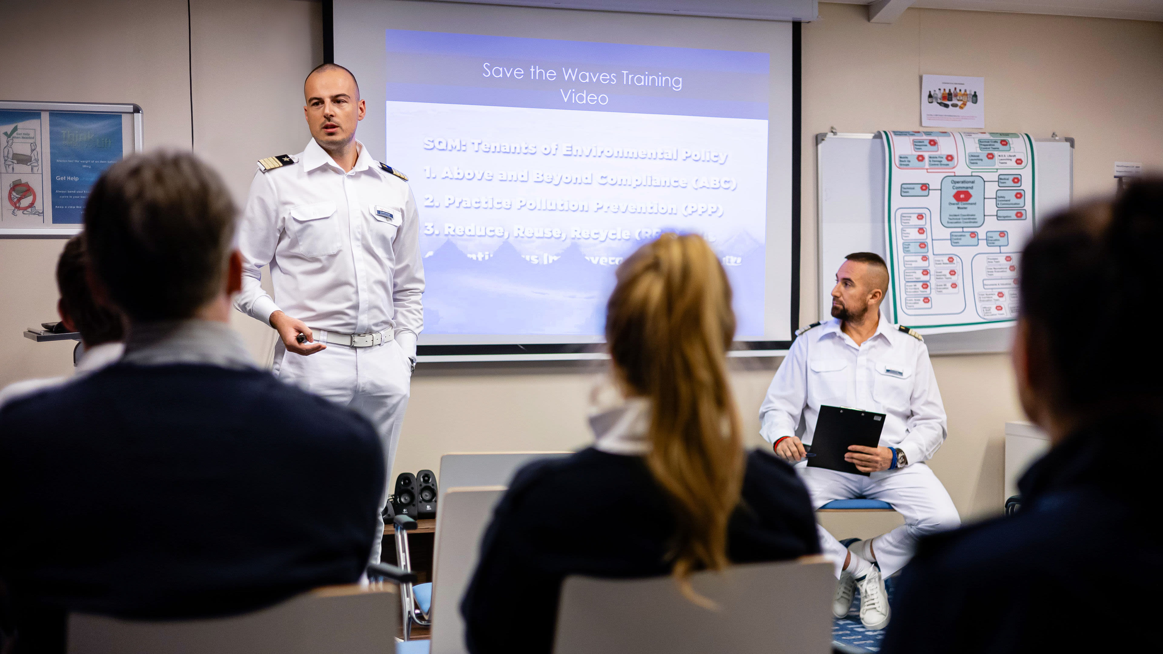 Mann in Uniform hält Vortrag vor Publikum, Präsentation im Hintergrund.