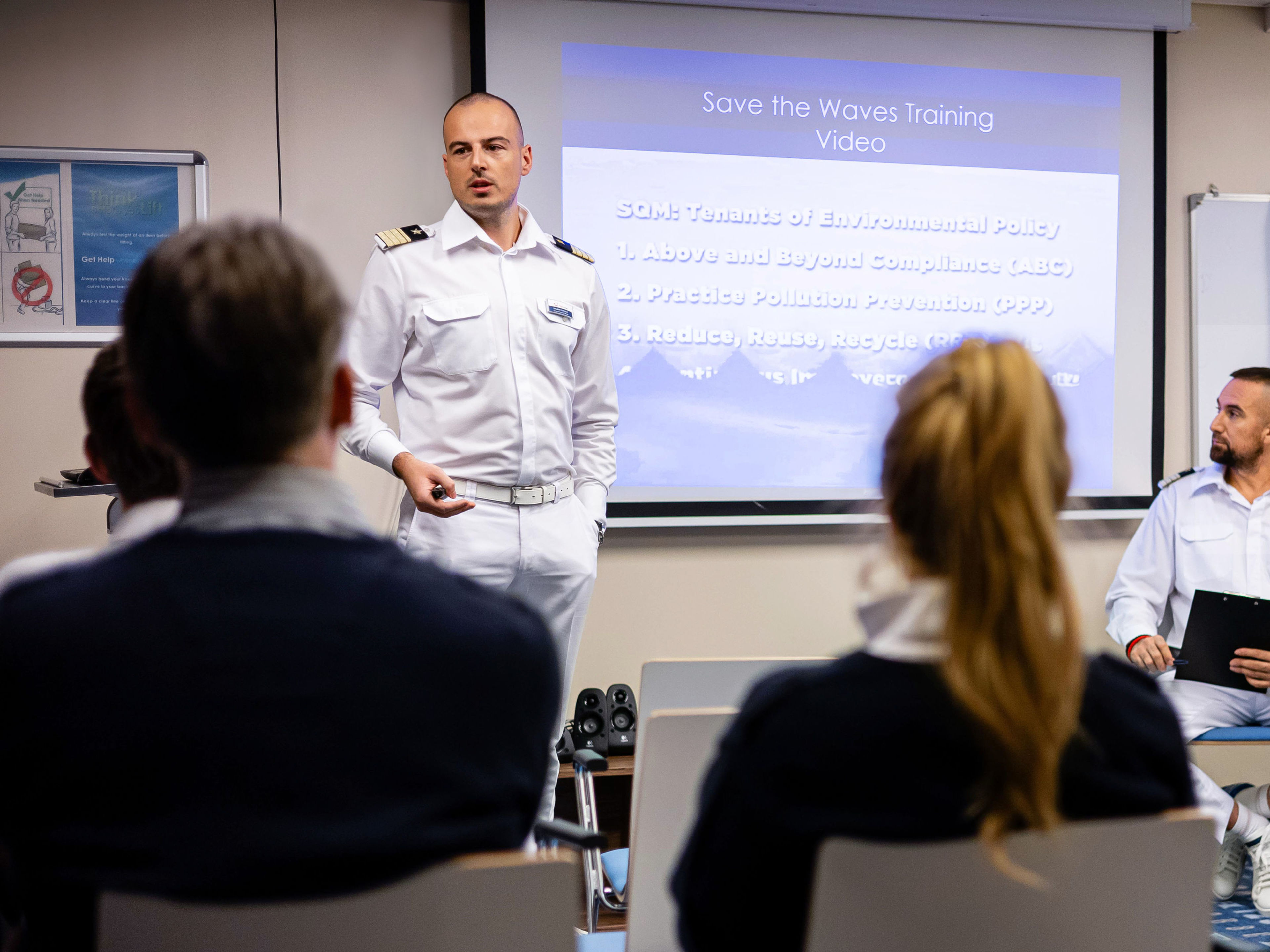 Umweltoffiziere sprechen mit der Crew Mann in Uniform hält Vortrag vor Publikum, Präsentation im Hintergrund.