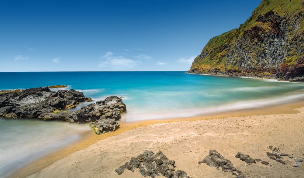 Sandstrand mit Felsen, blauem Meer und grüner Klippe unter klarem Himmel.