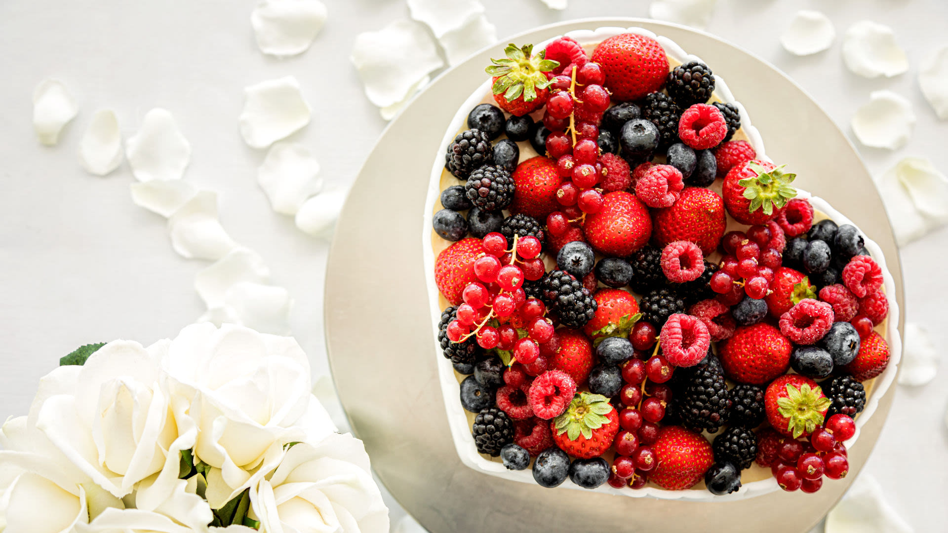 Herzförmige Beeren-Torte mit weißen Rosen und Blütenblättern.