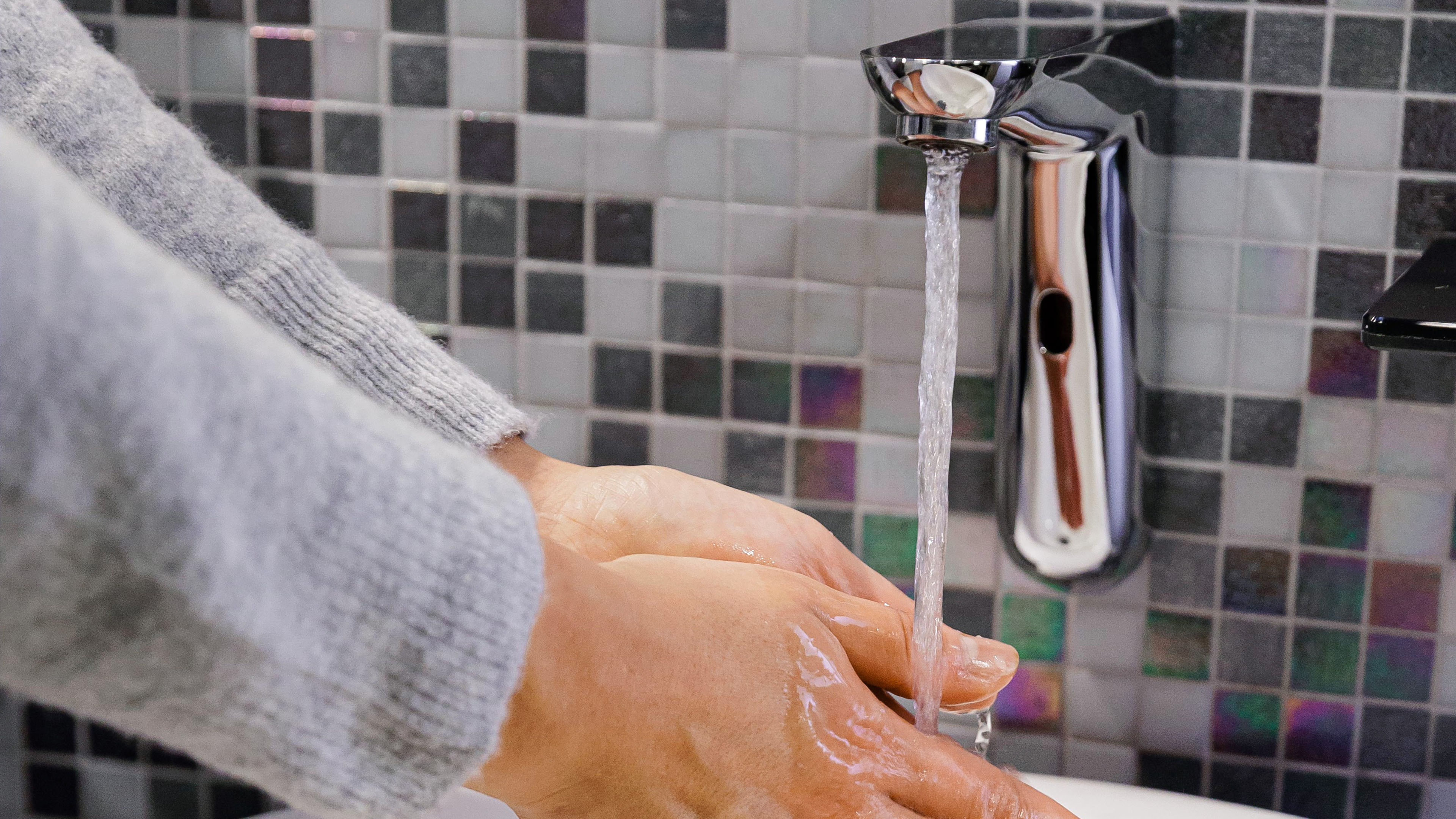 Hände waschen unter fließendem Wasser, graue Kacheln.