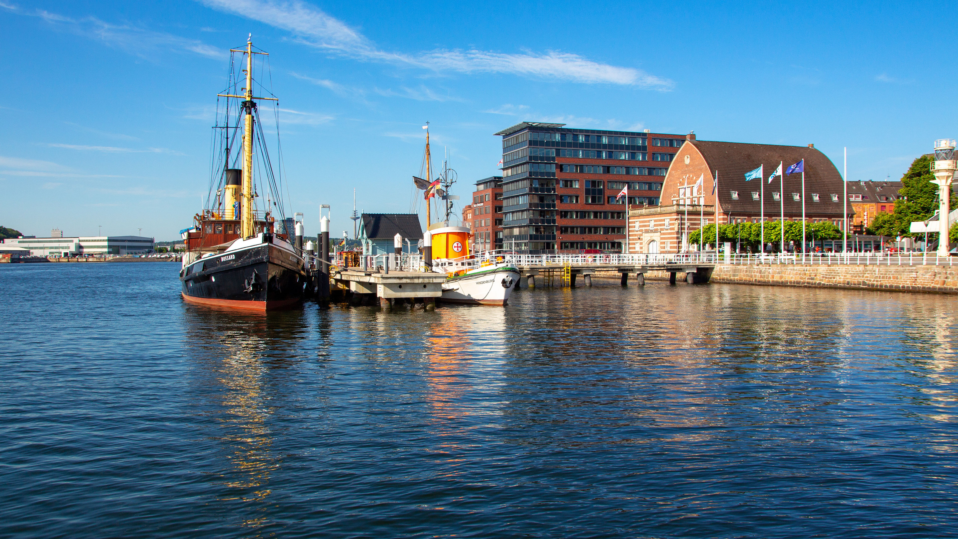 Hafen von Kiel mit historischen Schiffen und Gebäuden bei blauem Himmel.