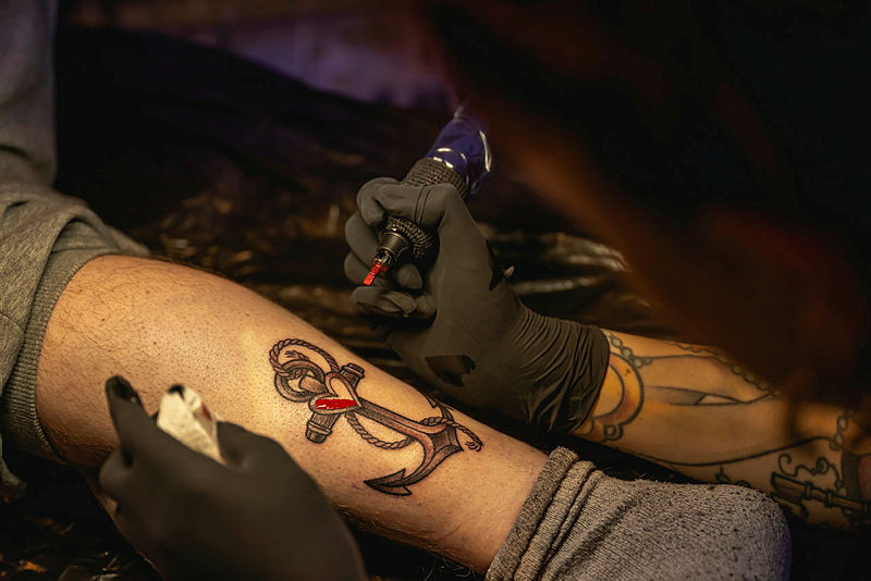 Tätowierer sticht Anker-Tattoo mit Herz auf Bein.