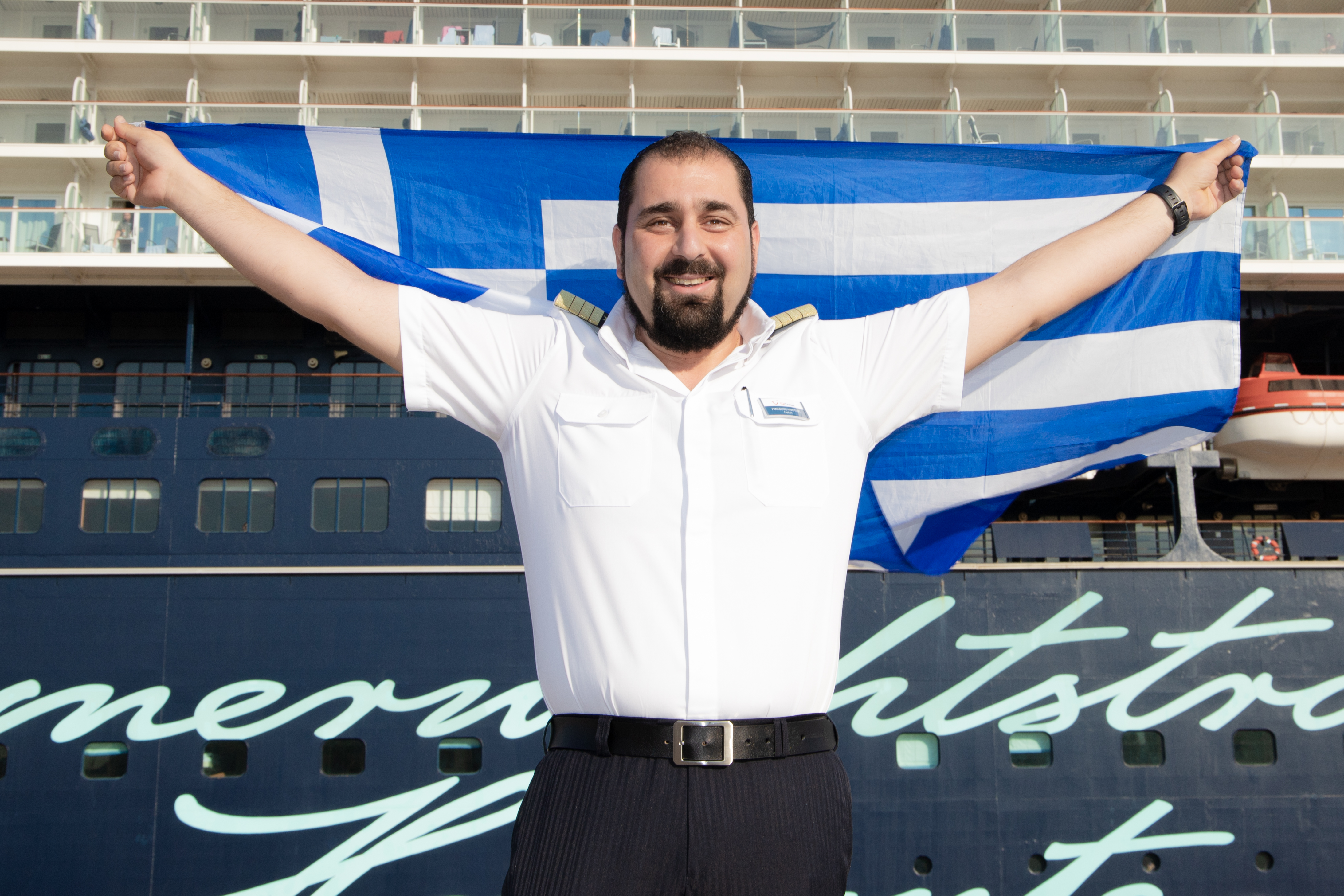 Mann in Uniform hält griechische Flagge vor Kreuzfahrtschiff der Mein Schiff® Flotte.