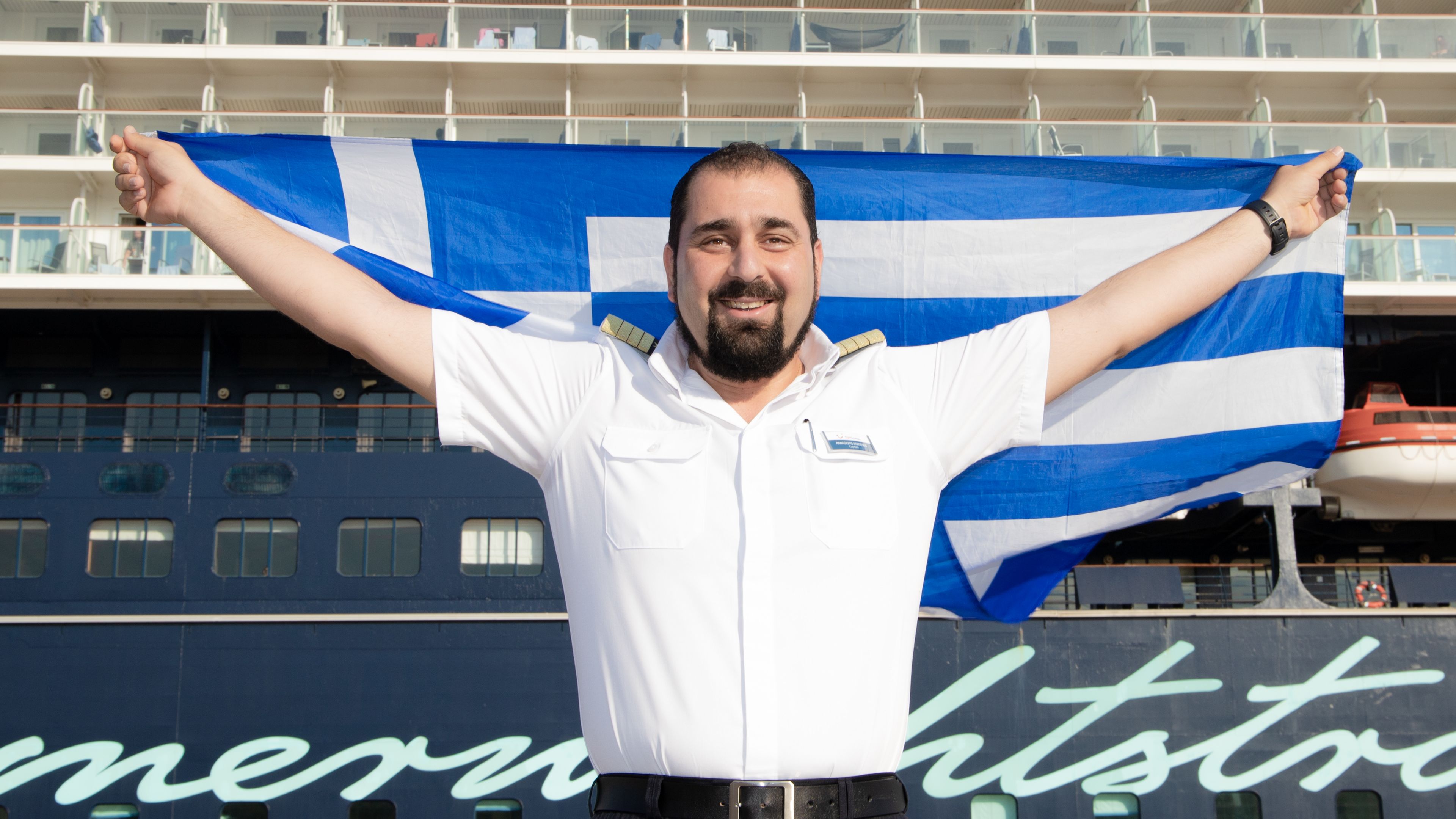 Panagiotis Varotsos Mann in Uniform hält griechische Flagge vor Kreuzfahrtschiff der Mein Schiff® Flotte.
