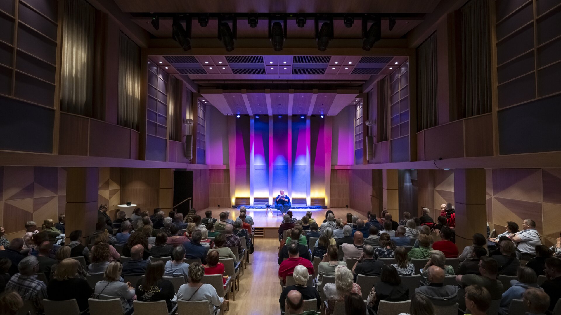 Publikum in einem modernen Konzertsaal, bunte Bühnenbeleuchtung.