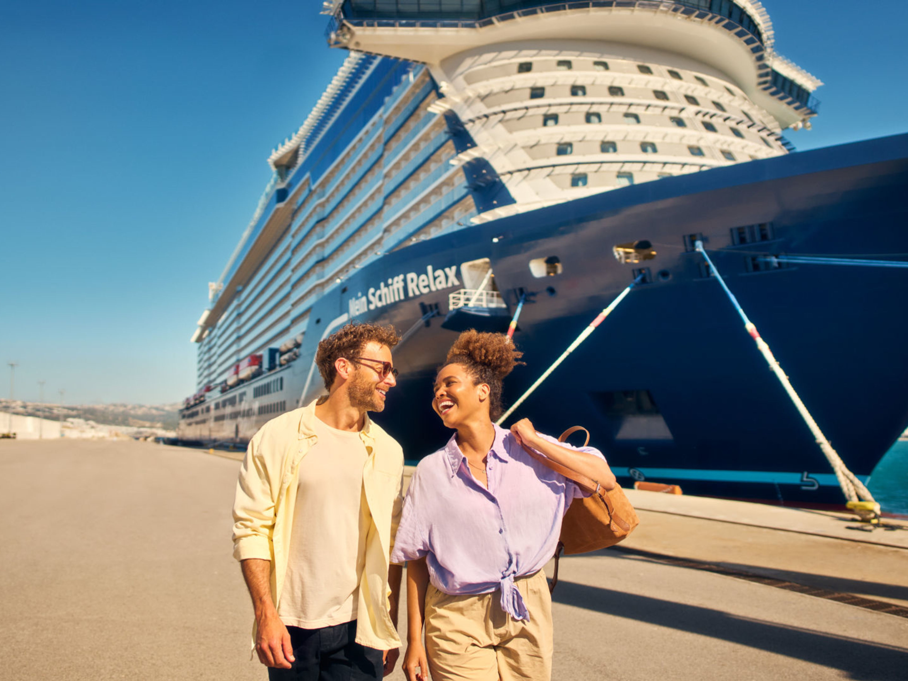Ein Pärchen spaziert am Anleger mit der Mein Schiff Relax im Hintergrund. Paar spaziert vor Kreuzfahrtschiff der Mein Schiff® Flotte, sonniger Tag.