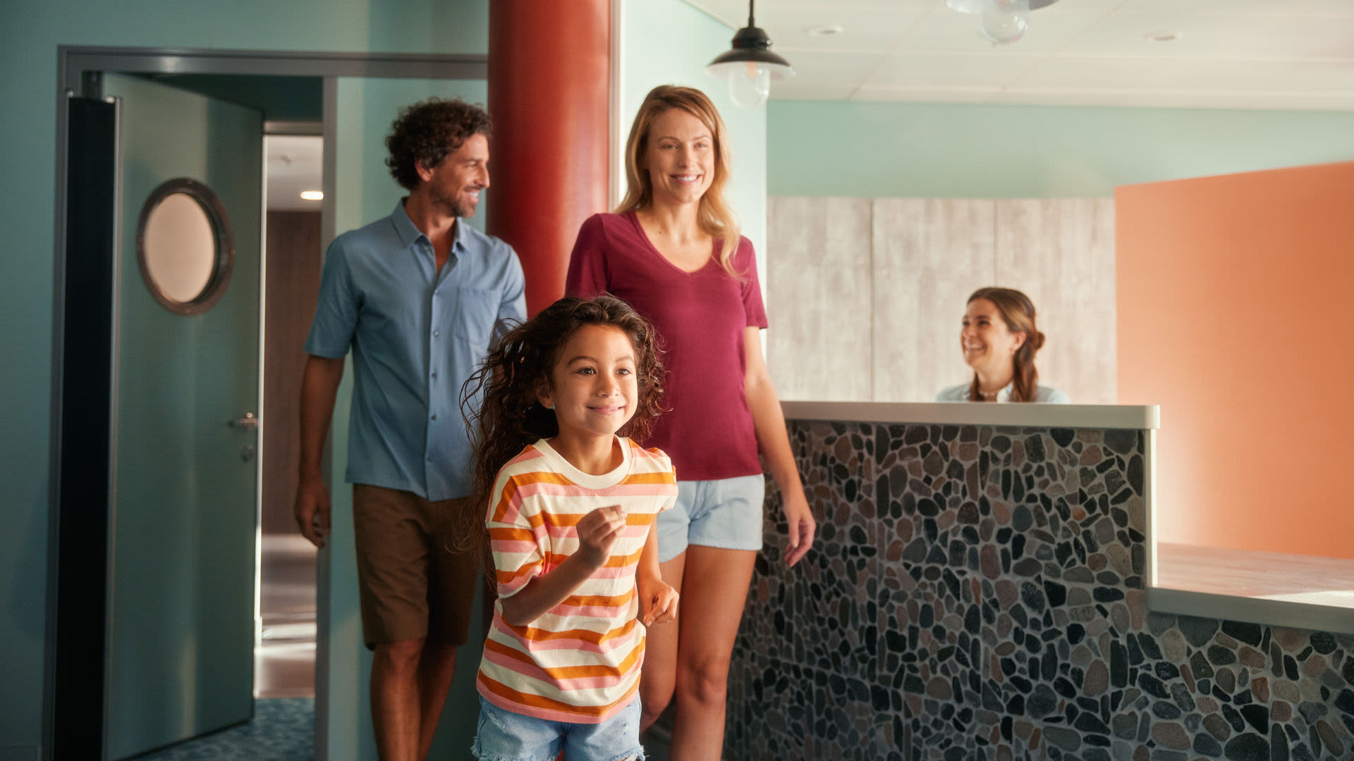 Familie betritt lächelnd Hotelrezeption, Empfangsdame begrüßt sie.