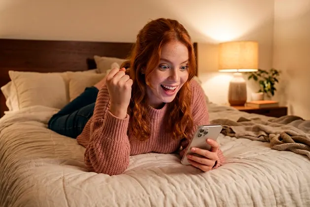 Foto de mujer joven acostada boca abajo en su cama mientras mira la pantalla de su celular mientras celebra con el puño de su mano izquiera arriba