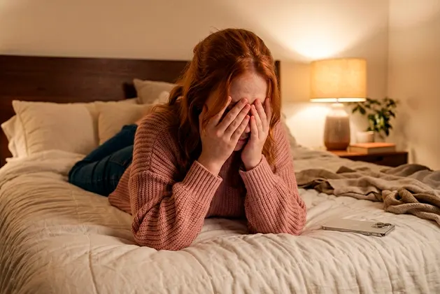 Foto de mujer joven acostada boca abajo en su cama mientras recuesta de manera desgonzada su cabeza en sus manos, tapando sus ojos