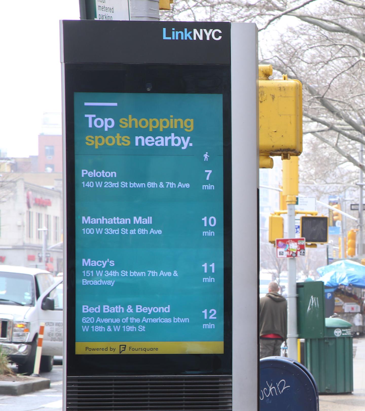 An Image of a digital board in NYC called "Link NYC". It is displaying a list of "Top Shopping spots nearby" which include Peloton, Manhattan Mall, Macy's and Bed Bath and Beyond with the prospective distance in minutes.