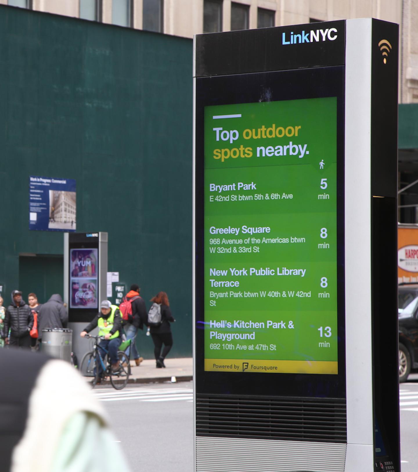 An Image of a digital board in NYC called "Link NYC". It is displaying a list of "Top Outdoor spots nearby" which include Bryant Park, Greeley Square, New York Public Library Terrace, and Hell's Kitchen Park and Playground with the prospective distance in minutes.