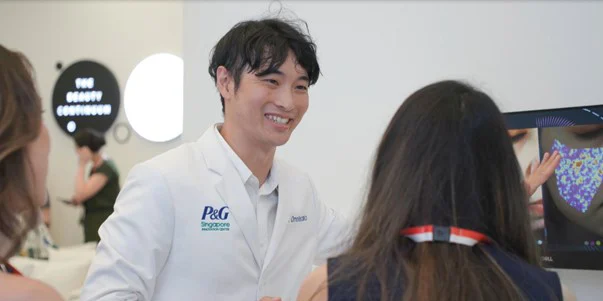 Zako-san, a young male Japanese scientist in a lab coat, gestures to a screen, helping a young woman understand her Y-age skin analysis results