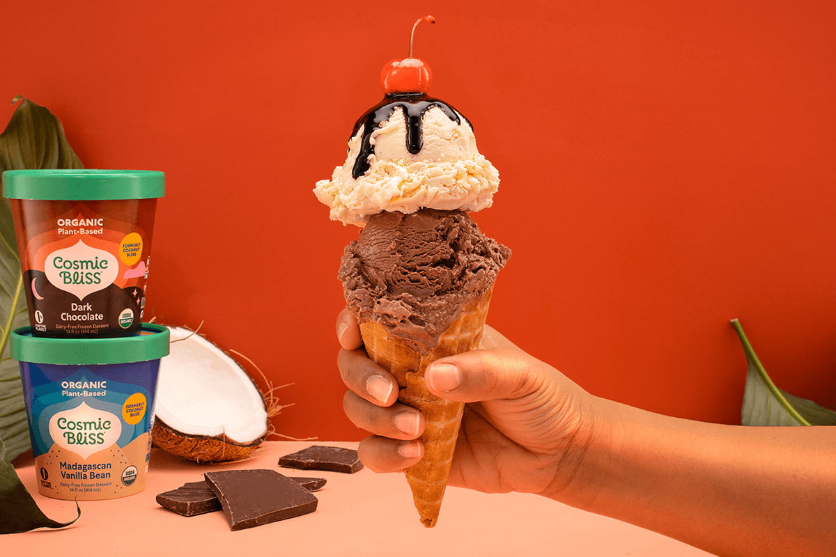 hand holding an ice cream cone with one chocolate scoop, one vanilla scoop chocolate sauce and a cherry on top. In the background there are also 2 pints of ice cream stacked on top of one another and a half of a coconut. 