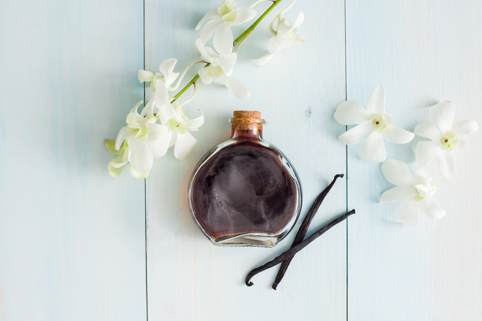 Background of white washed wood with glass bottle of vanilla extract surrounded by vanilla flowers and beans