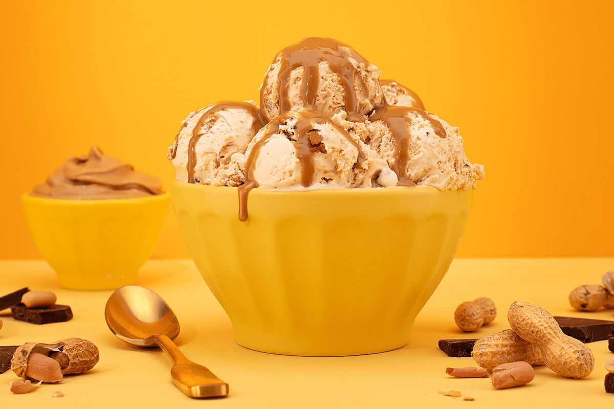a yellow bowl overflowing with scoops of indulgence peanut butter ice cream and surrounded by peanuts and chocolate chunks
