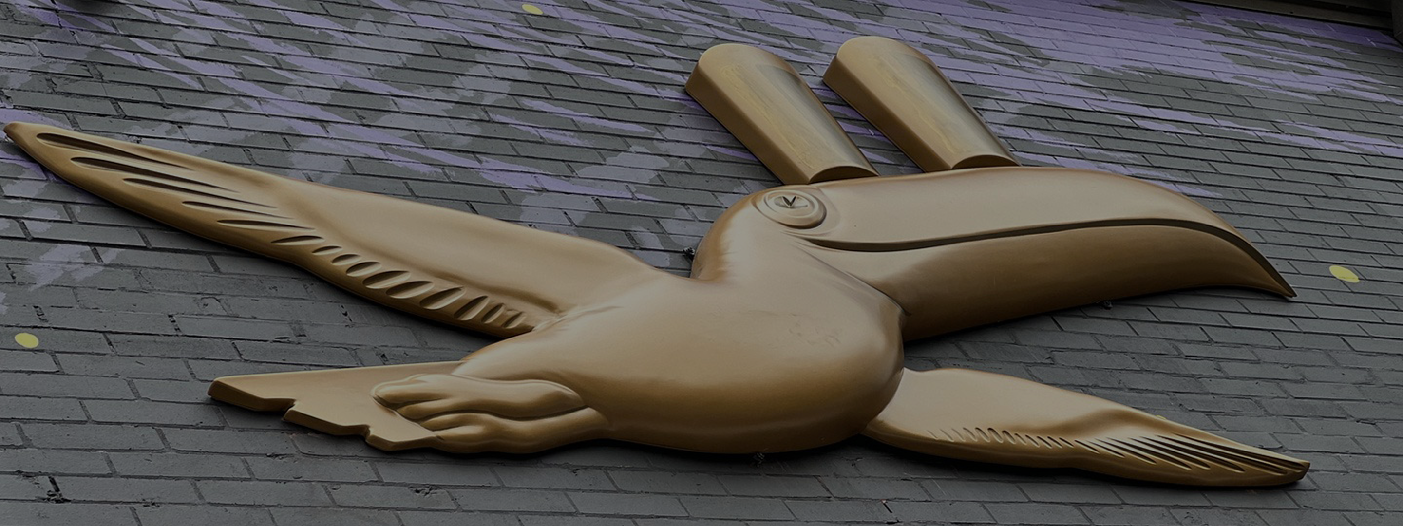 Gold-colored sculpture of a stylized bird with two cylindrical shapes on its head, mounted on a gray brick wall