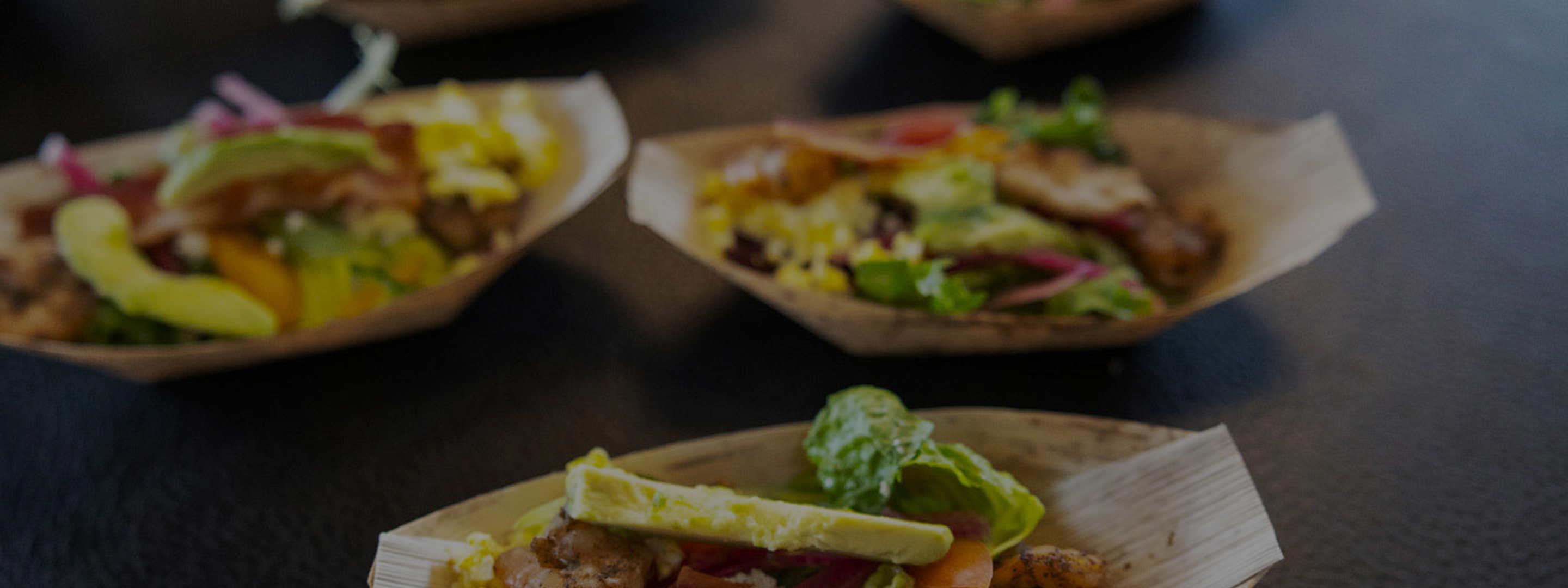 Several small trays filled with assorted food, including avocado, greens, and grilled items, arranged on a dark surface.