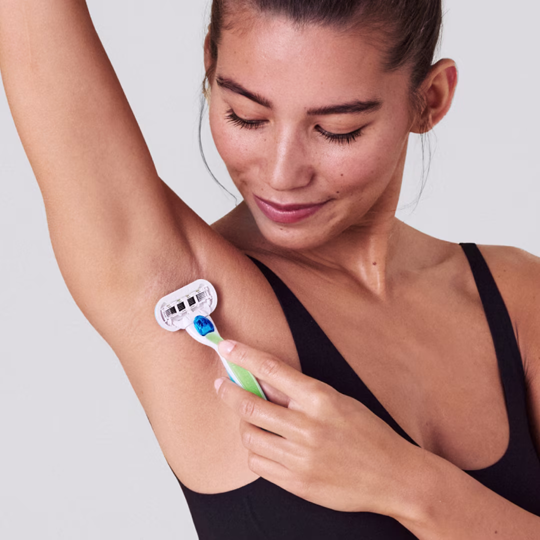 Woman in a black top shaving her underarm with a blue razor, looking down and smiling.