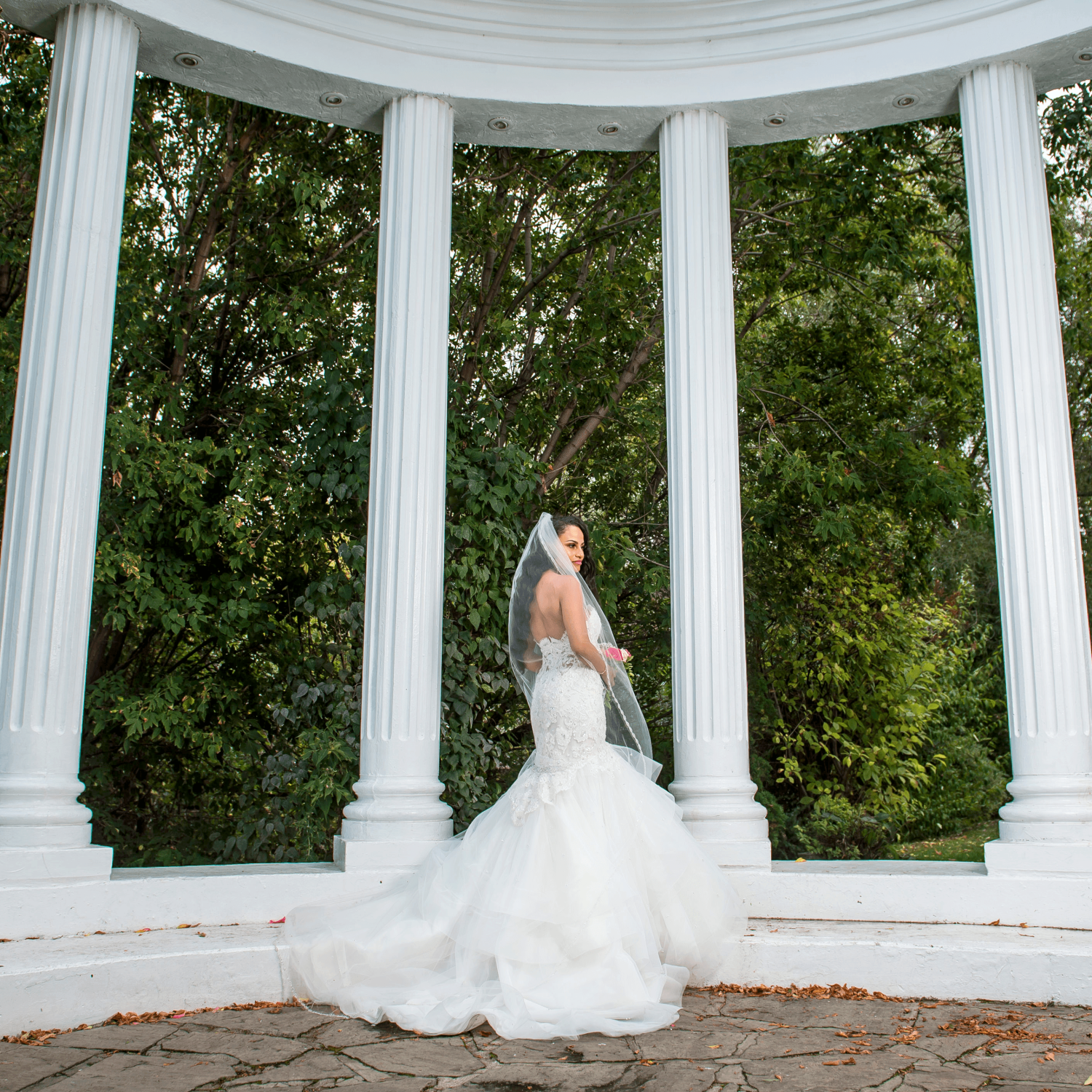 A bride standing in the Paradise Banquet Halls garden venue's columned area with lush, green trees in the background.