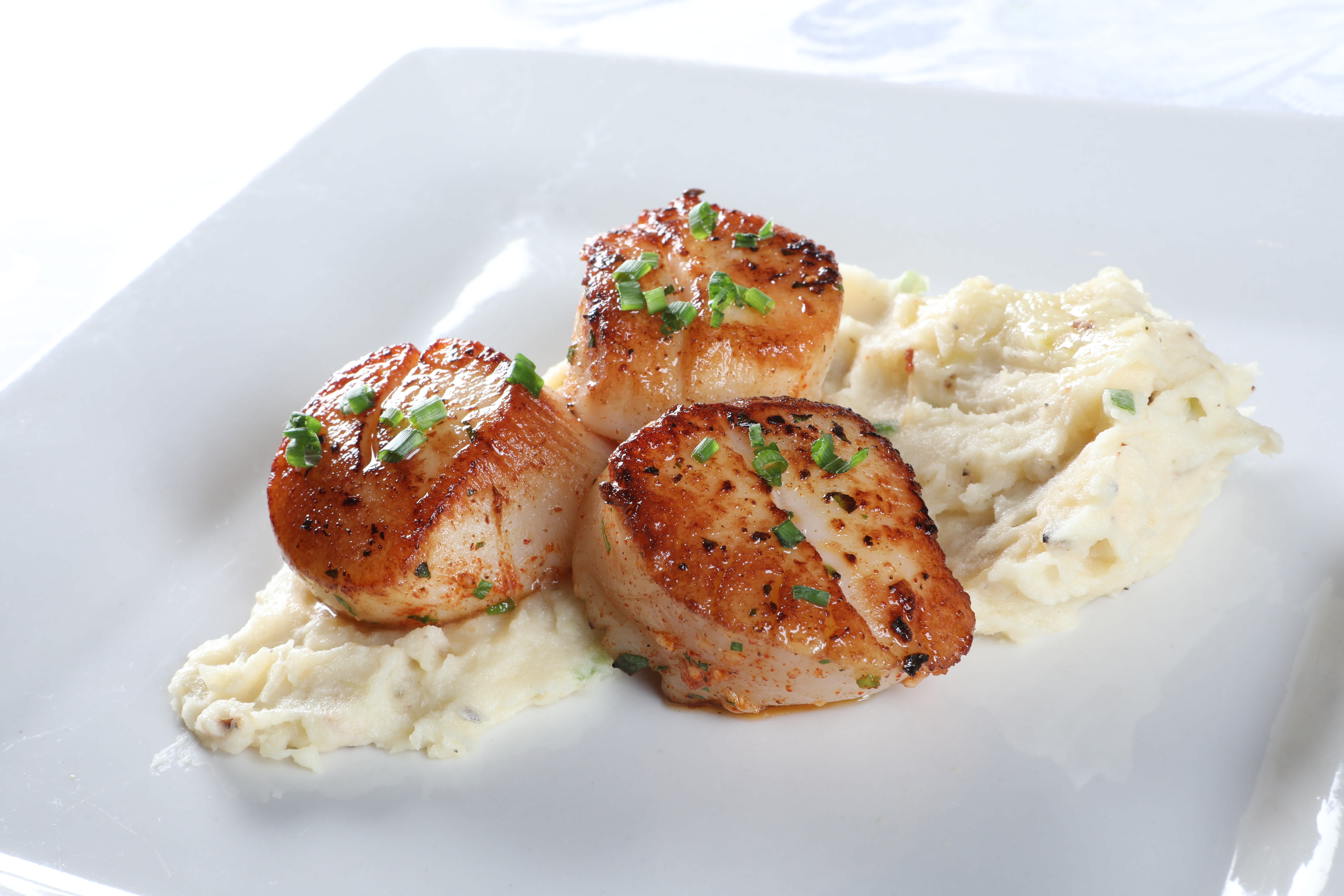 A plate of seafood with Potato coulis, greens, and Chives is served on a white plate which is placed on a marble countertop.