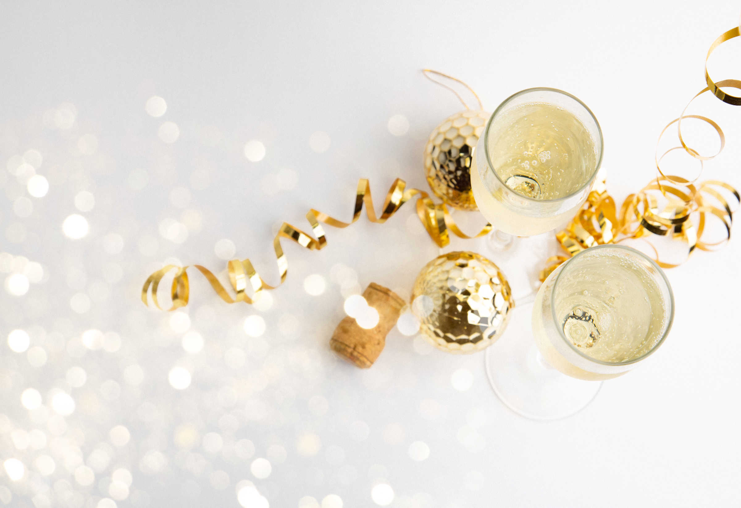 A couple of champagne glasses half full of champagne with curled, golden streamers and two golden ball ornaments with bokeh sparkles to the left.