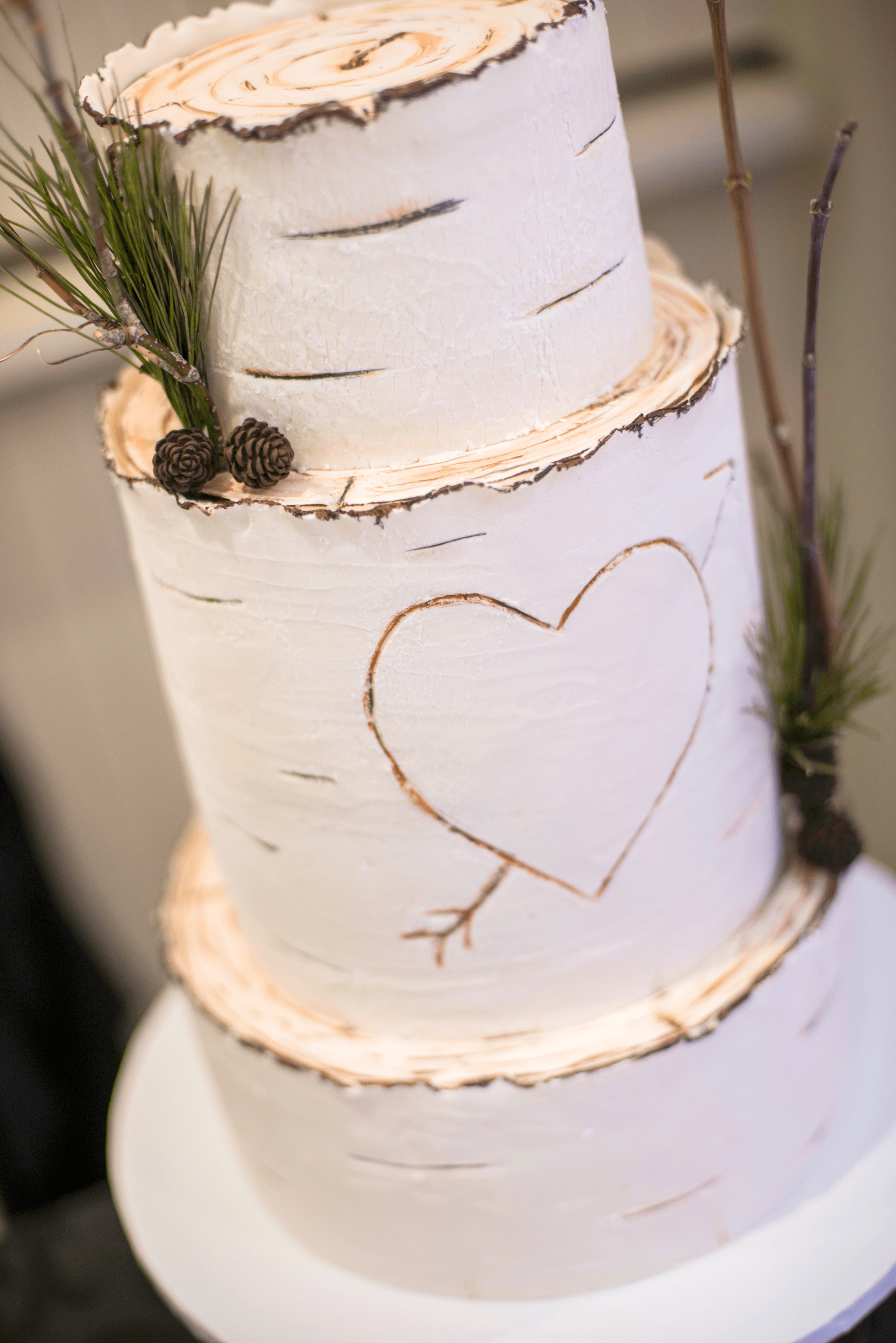 A three-tiered white cake resembling birch bark has pine springs and cones as well as a heart with an arrow through it carved into the front. Made by the culinary team at Paradise Banquet Halls.