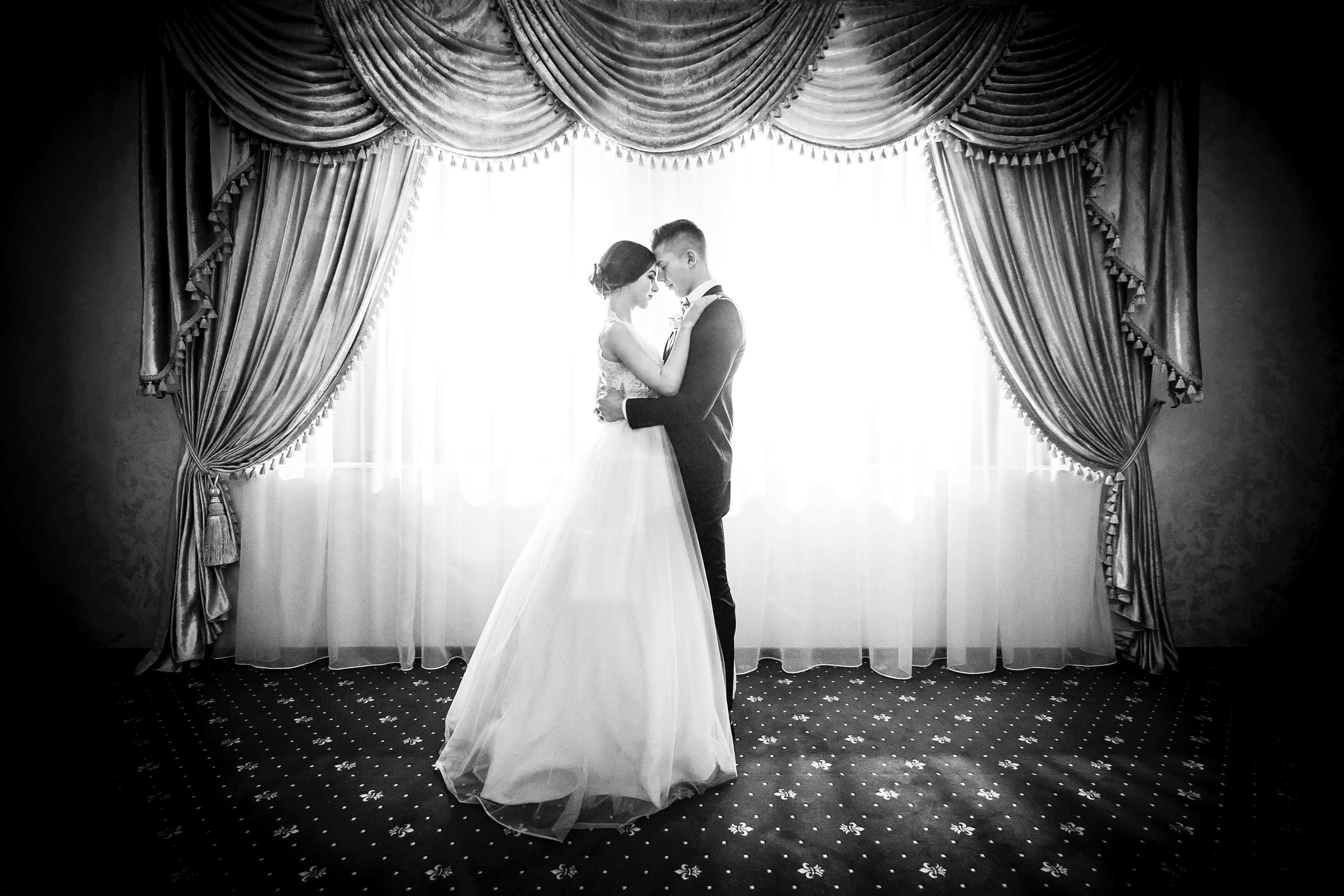 The wedding couple embracing in front of a curtained window in black and white.