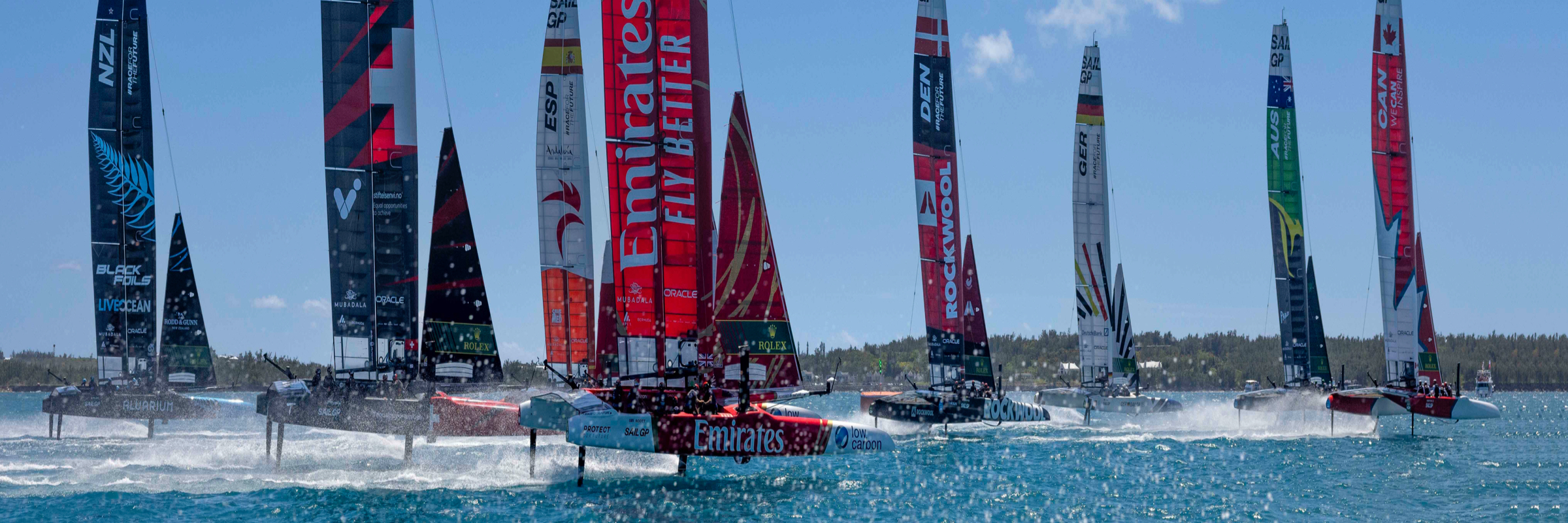 A high-speed sailboat on foils rides above the water, with a red and black hull featuring a Swiss flag. Five crew members in helmets and life jackets are on board, actively maneuvering. The sail has the text "#backthebid" and another boat is visible in the background.