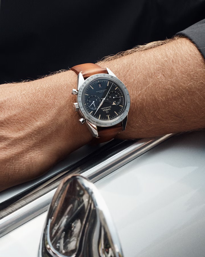 A close-up image of a person's wrist wearing a luxury chronograph watch with a brown leather strap. The watch face is black with multiple sub-dials and silver hour markers. The person's arm is resting on a car, with part of its chrome side mirror visible.