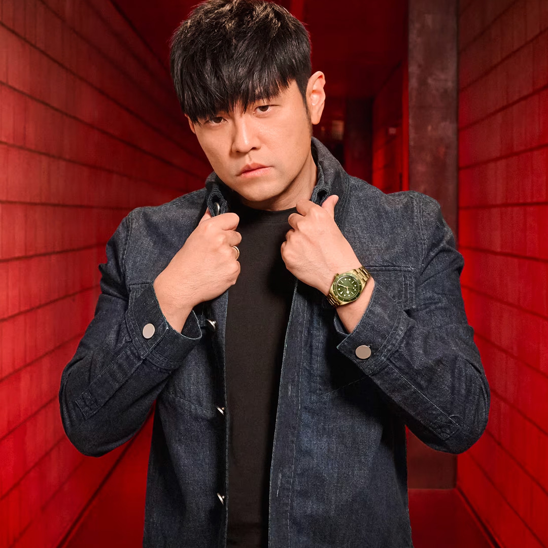 A person with straight, dark hair wearing a dark denim jacket and a gold watch stands in a dimly lit corridor with red walls, holding the collar of the jacket with both hands and looking directly at the camera.