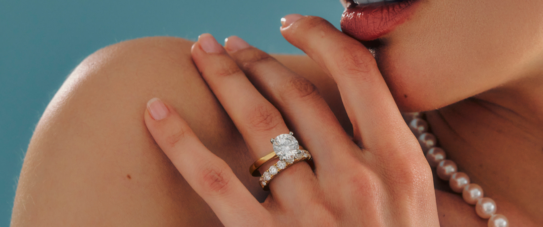 A close-up of a person's hands gently resting on a surface. The left hand features a gold ring set with a prominent, sparkling diamond and a wedding band adorned with multiple smaller diamonds. The person's nails are neatly manicured with a natural look.