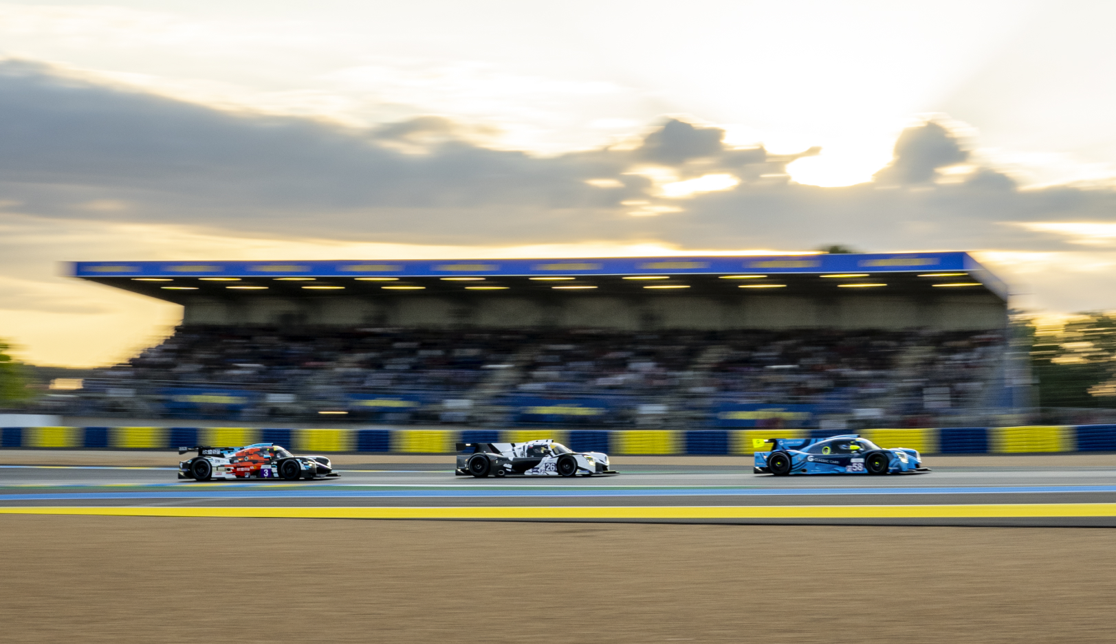 A group of race cars speed down a track under a green Rolex banner. The electronic sign above the track reads "FLAG START" and "1 LAP". Spectators and trackside structures are visible in the background. The scene is dynamic and intense.