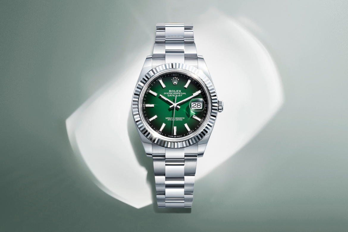 A luxury stainless steel wristwatch with a green dial and fluted bezel is displayed on a light green and white background with a honeycomb pattern. The watch features a date window and a metal link bracelet.