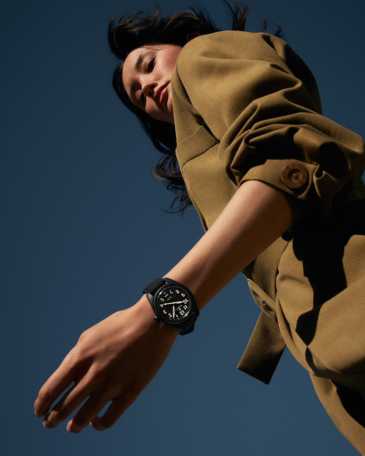 Low-angle view of a person wearing a tan jacket, seen from below against a dark blue sky. The person's wrist, adorned with a large black watch, is prominently in the foreground, positioned above their head, creating a dramatic and dynamic composition.
